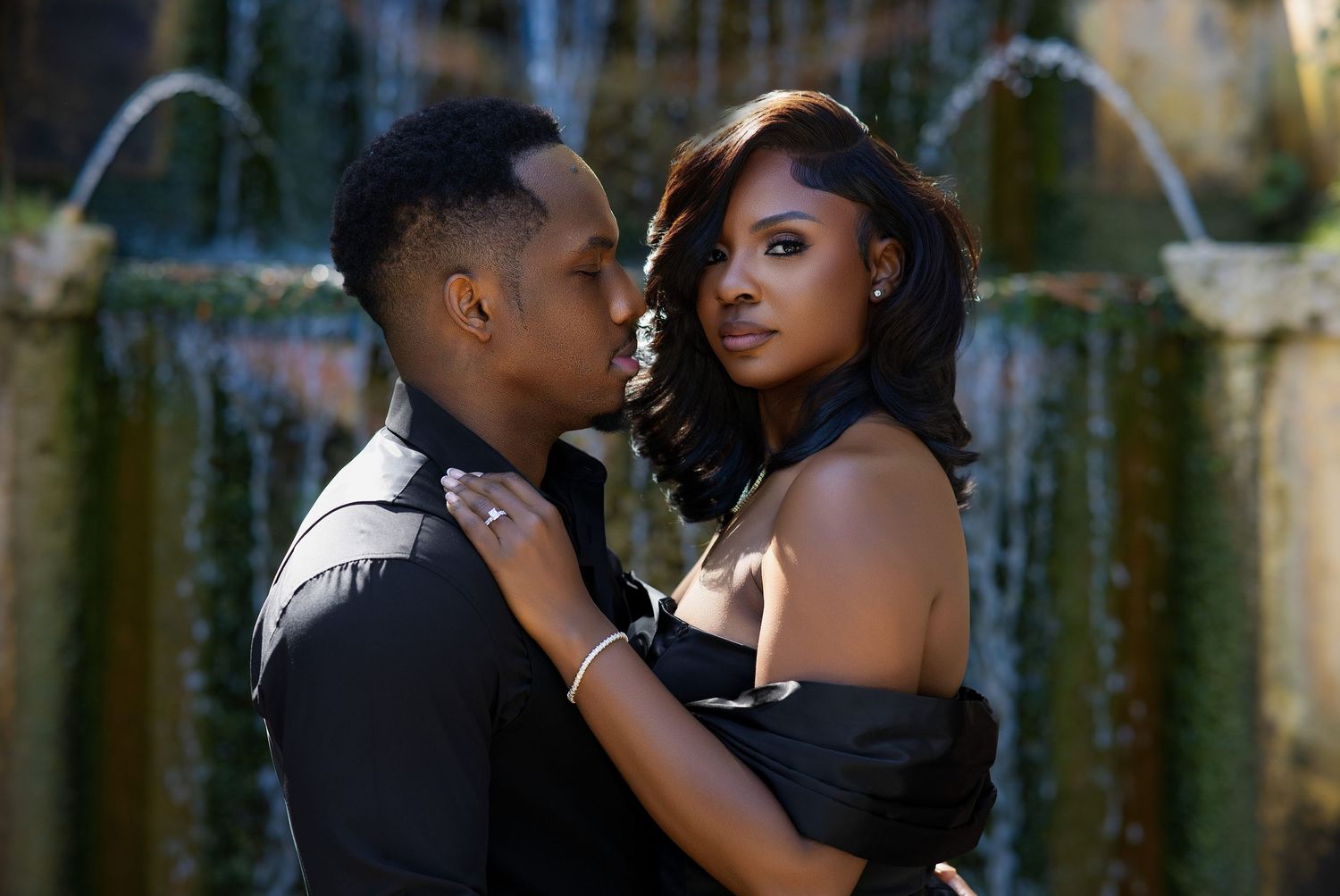 Romantic couple in black outfits posing intimately in front of a stone fountain.