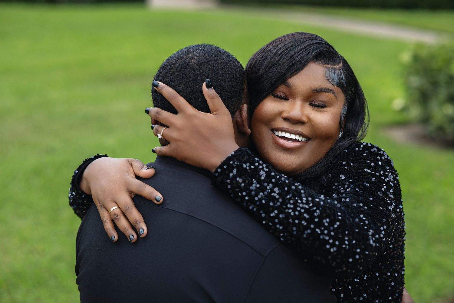 Smiling woman in black sequin gown joyfully embracing her partner outdoors on a green lawn.