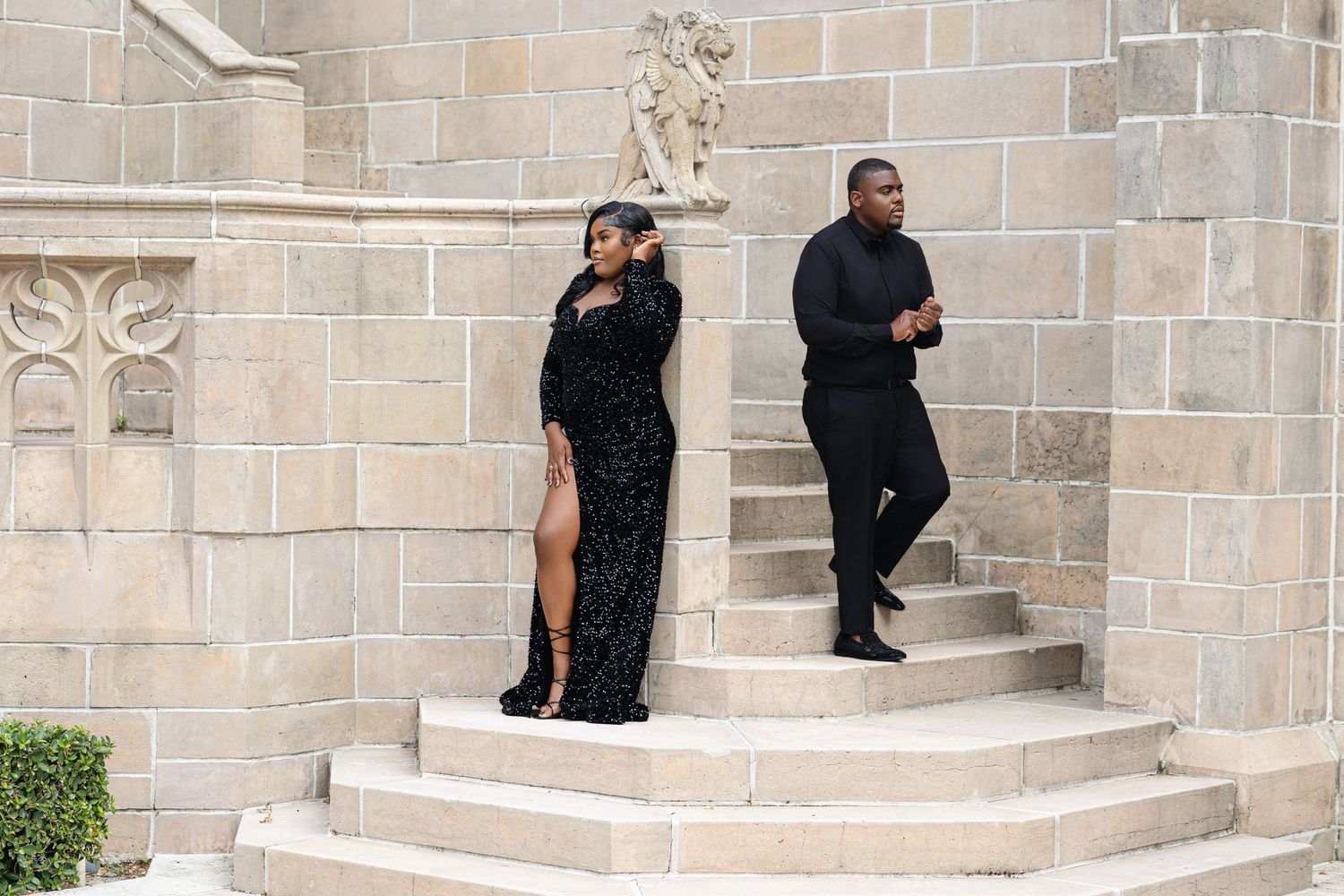 Couple in elegant black attire posing separately on stone steps of a historic Gothic stone building.