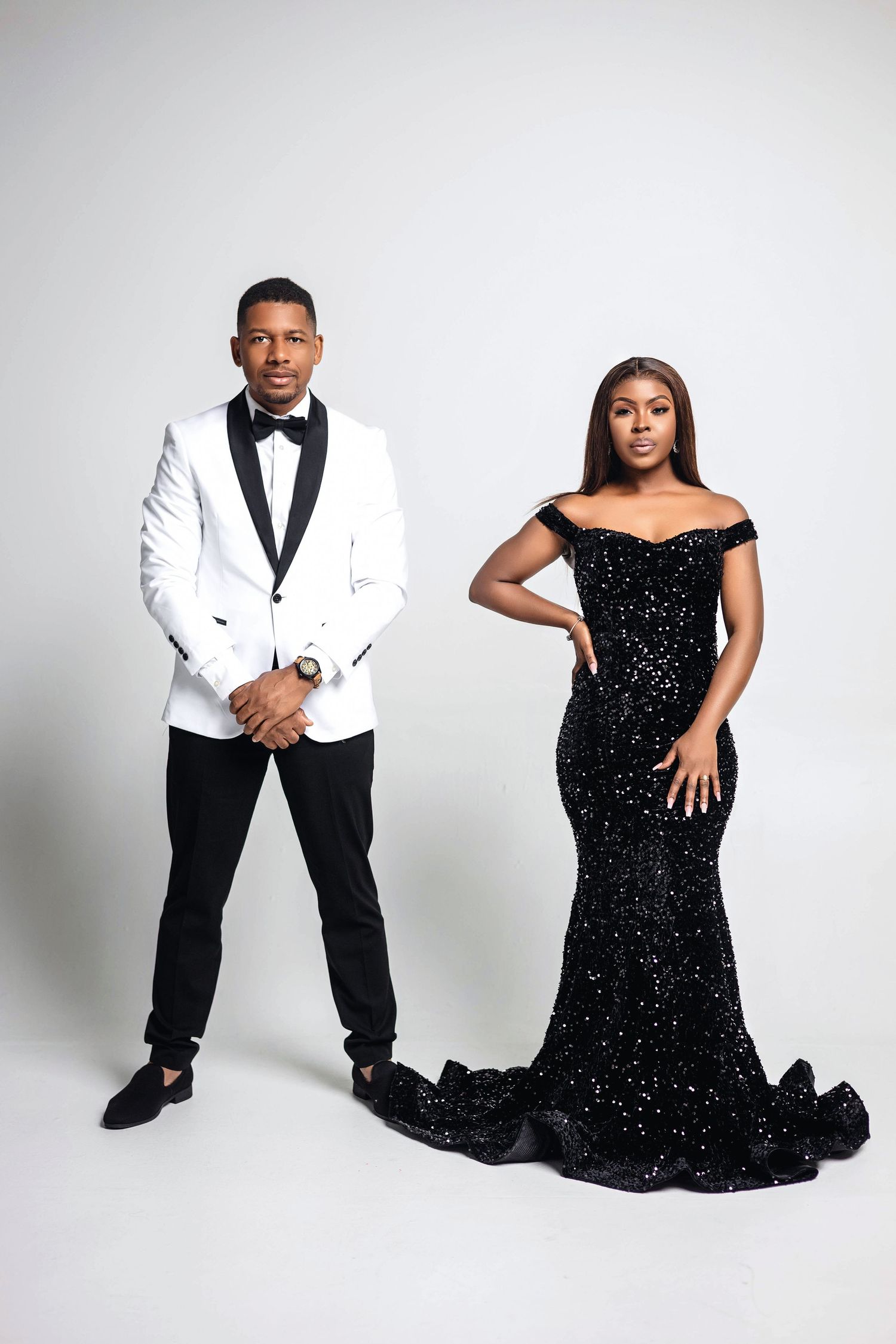 Couple posing on white backdrop, man in white tuxedo jacket, woman in sparkling black off-shoulder mermaid gown.