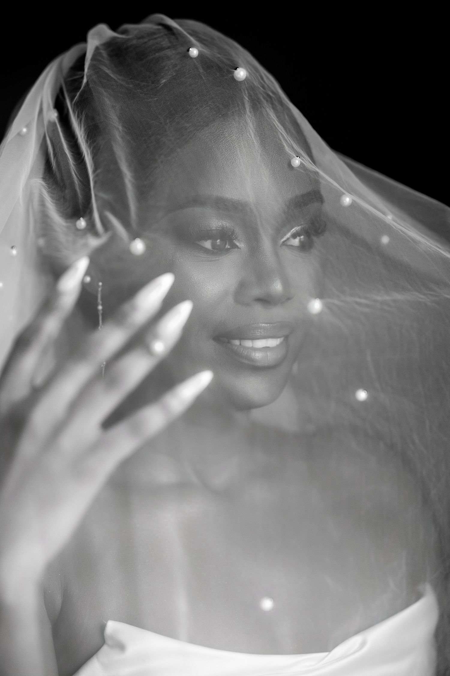 Black and white portrait of a bride wearing a pearl-adorned veil, smiling softly with manicured nails visible.
