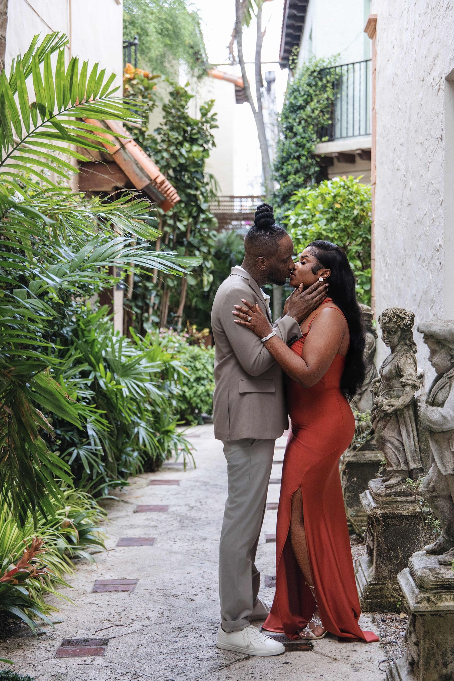 A Black couple shares a romantic moment in a lush garden alleyway adorned with stone sculptures.