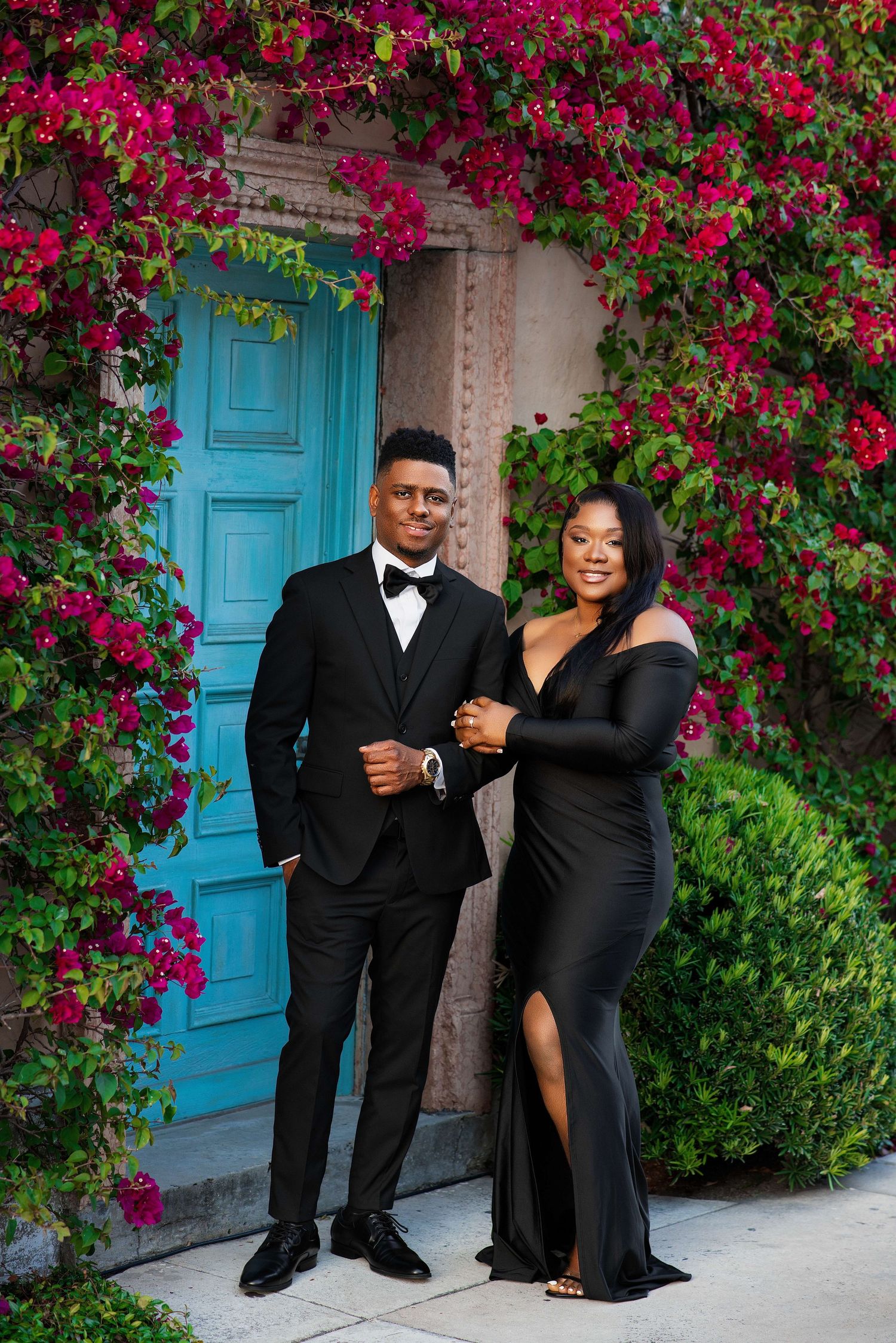 A couple in coordinating black formal wear pose before a vibrant bougainvillea-covered wall with a teal door.