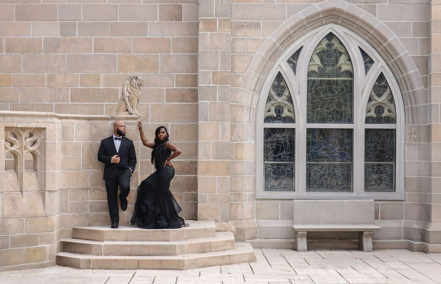 Elegant couple in black formal wear standing beside gothic stone window and lion sculpture outdoors.