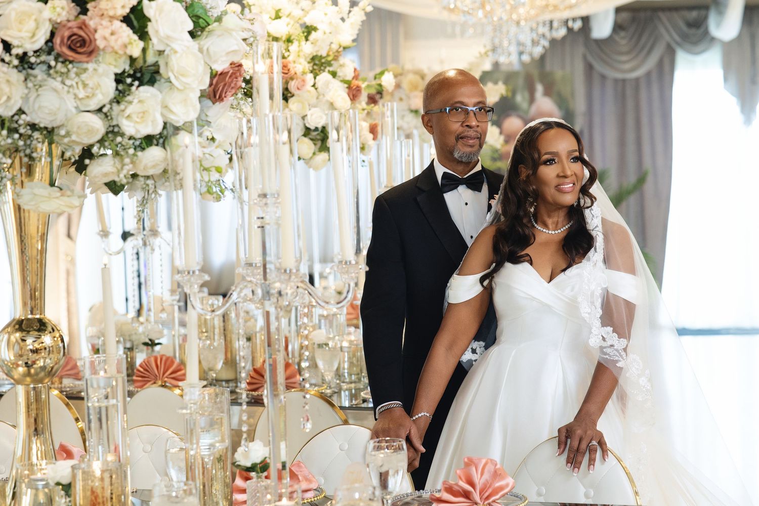Elegant bride and groom pose together at a luxurious wedding reception with tall floral arrangements and gold candelabras.