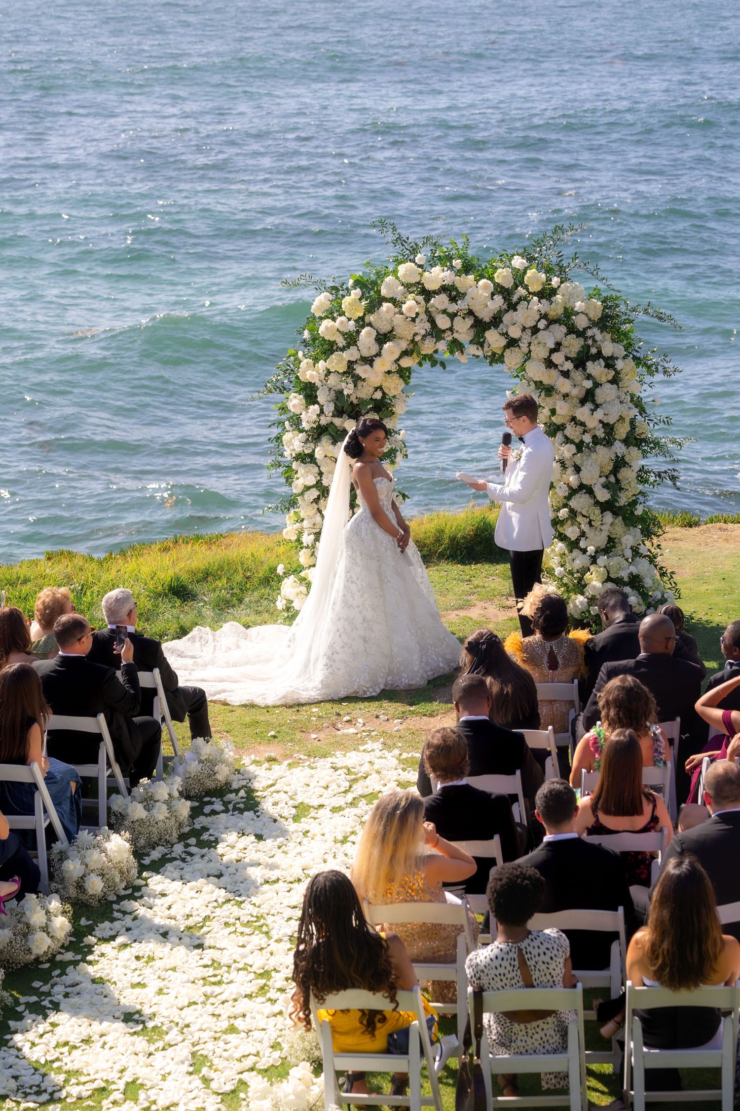 Outdoor wedding ceremony by the ocean with couple at floral arch, white roses, and seated guests on white chairs.
