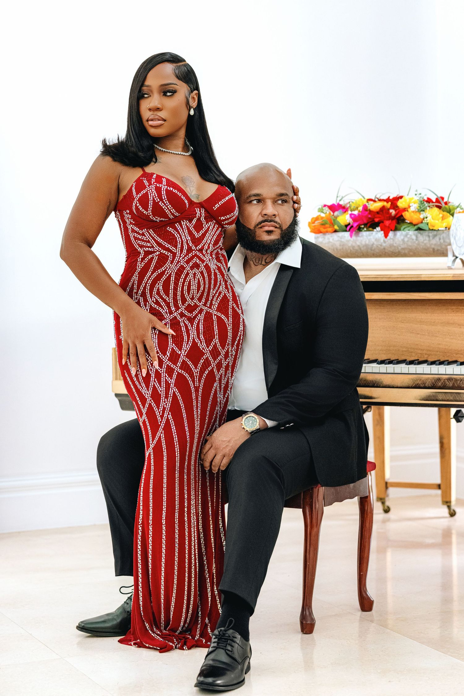 Couple in elegant formalwear poses by a grand piano, woman in stunning red beaded gown, man in black suit.