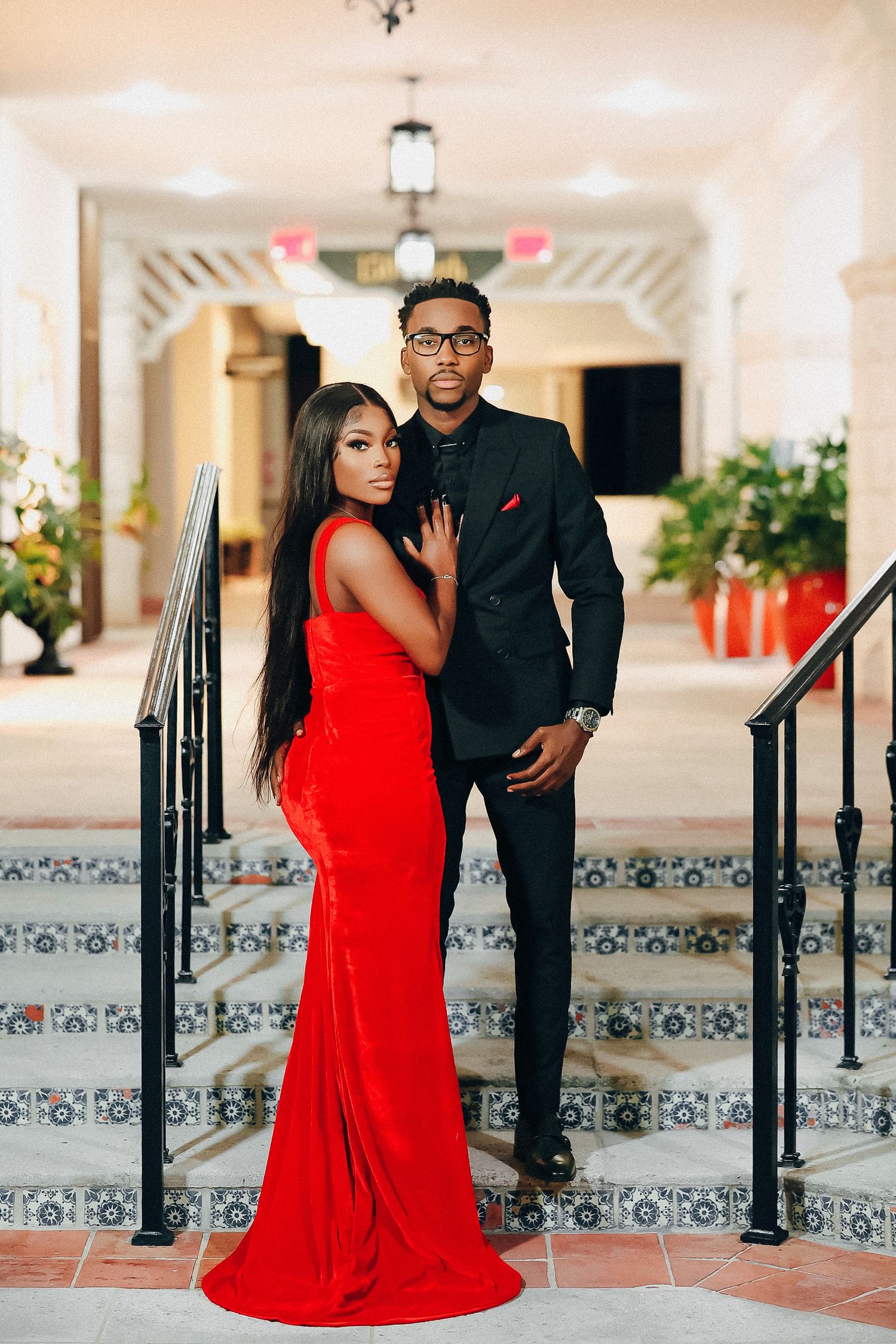 Elegant couple in red gown and black suit posing on decorative tiled steps in a grand interior hallway.
