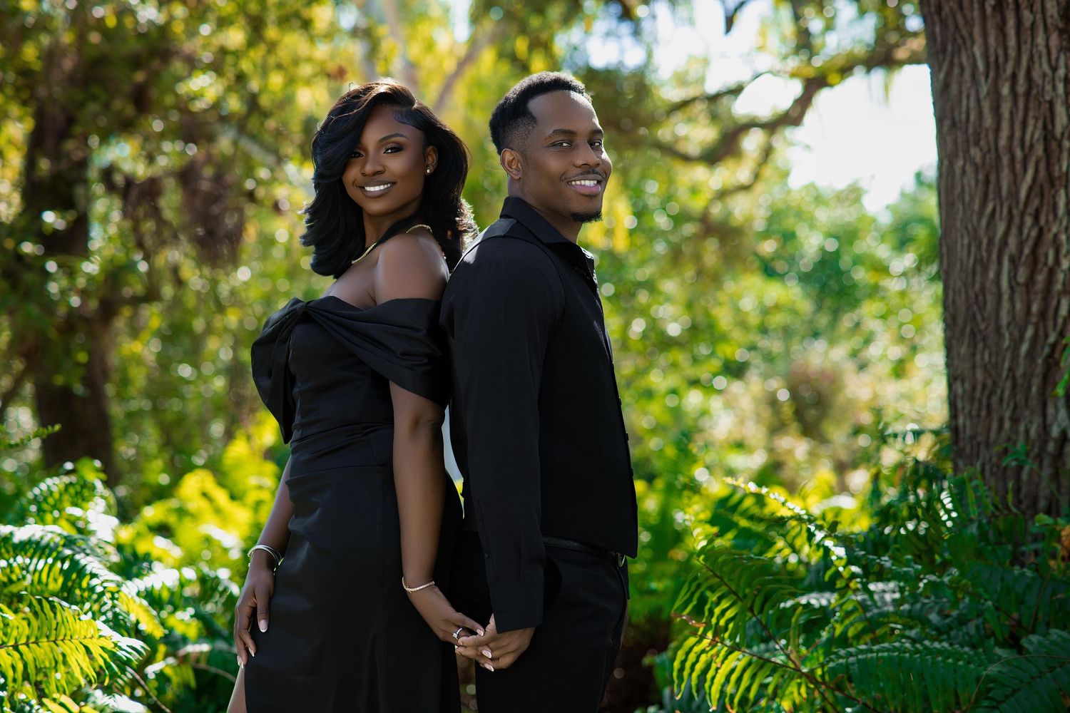 Black couple in matching black formal wear holding hands and smiling in lush green garden setting.