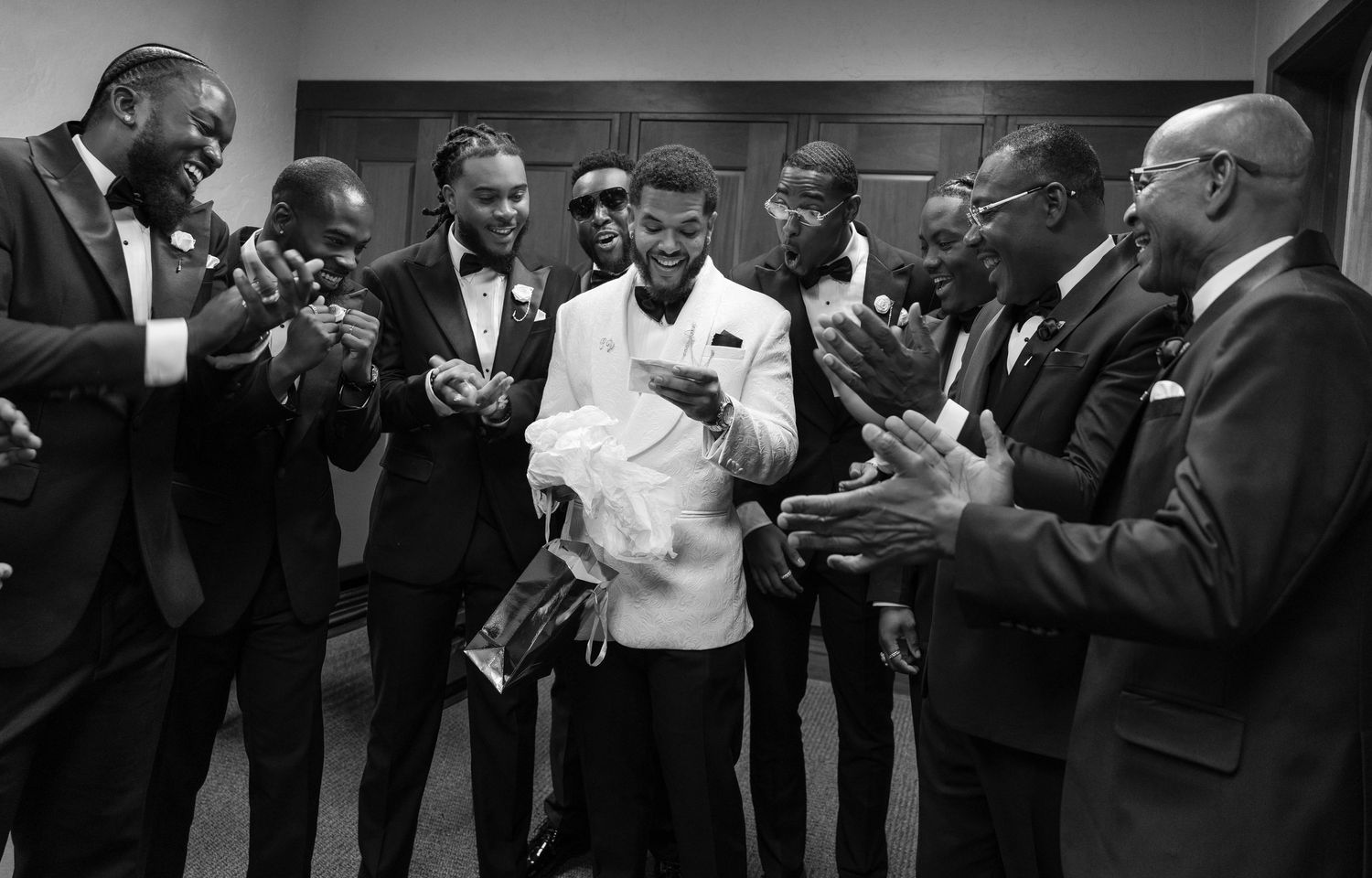 Black and white photo of a groom in white jacket surrounded by applauding groomsmen in tuxedos.