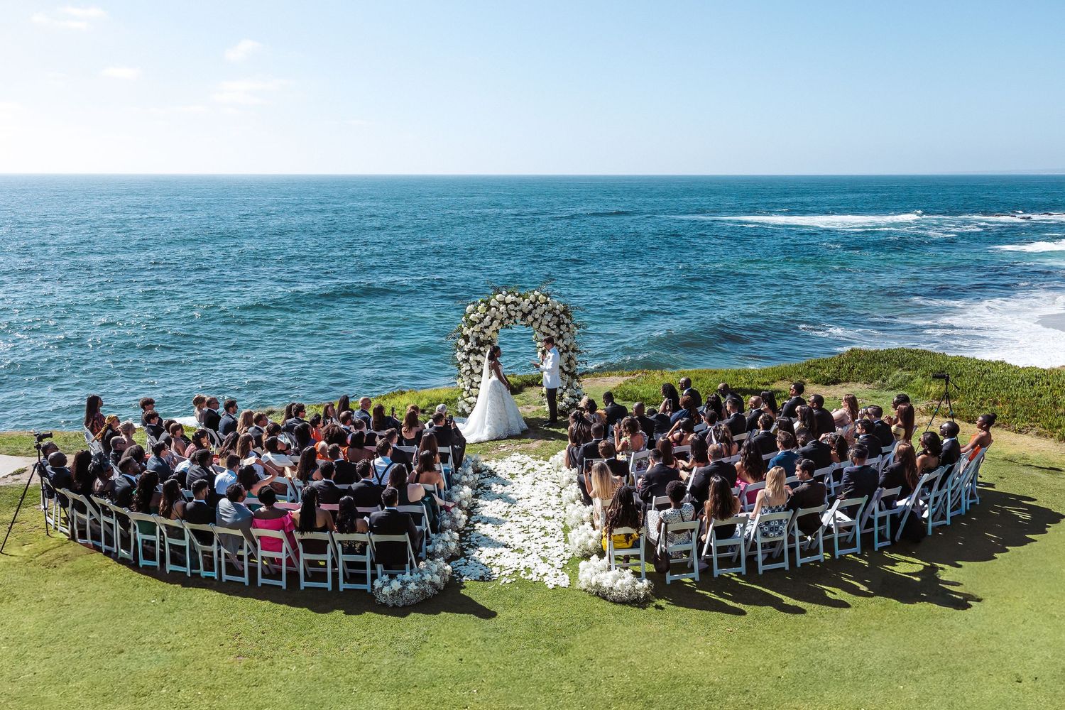 Outdoor wedding ceremony on ocean cliffside with floral arch, white chairs, and guests seated on lush green lawn.