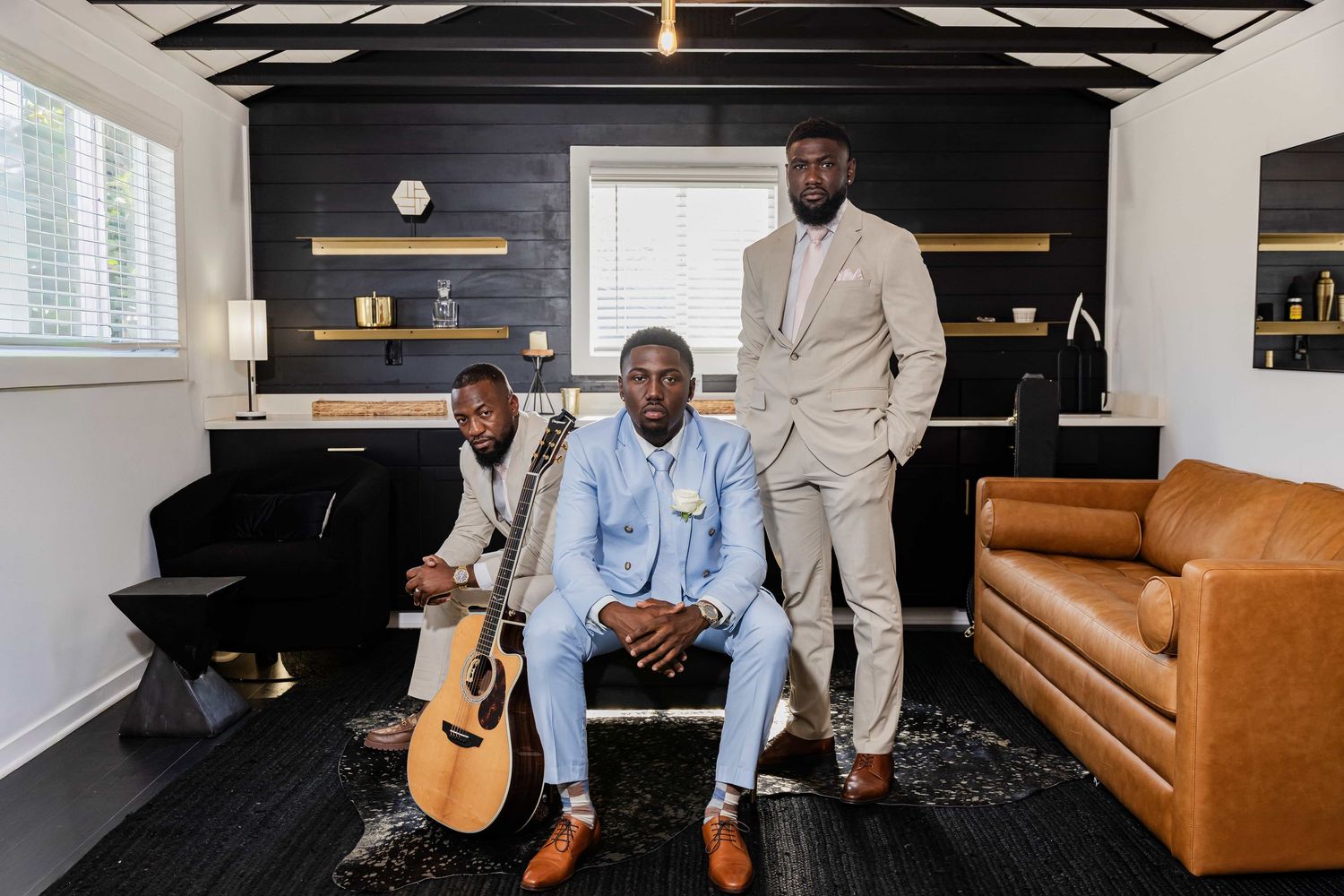 Three well-dressed men pose in a modern office, one holding an acoustic guitar, surrounded by stylish furniture.