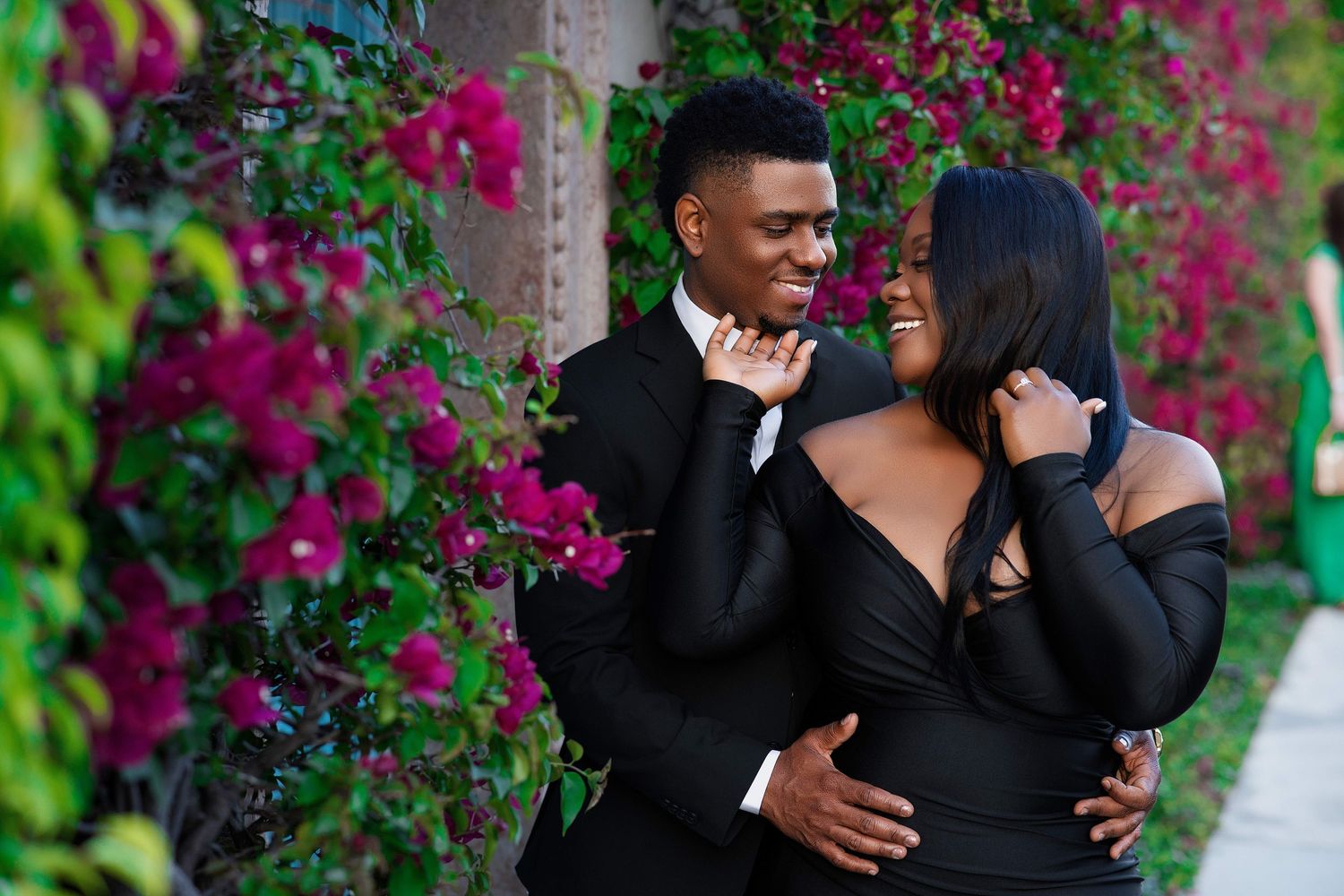 A joyful couple in elegant black attire share a playful, loving moment surrounded by blooming bougainvillea.