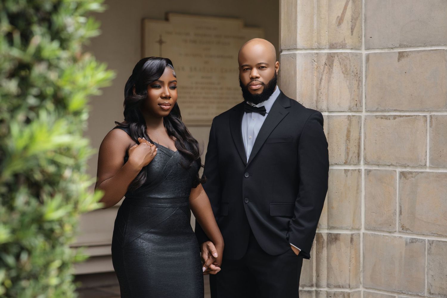 Elegant couple in black formal attire holding hands beside stone wall with lush greenery.