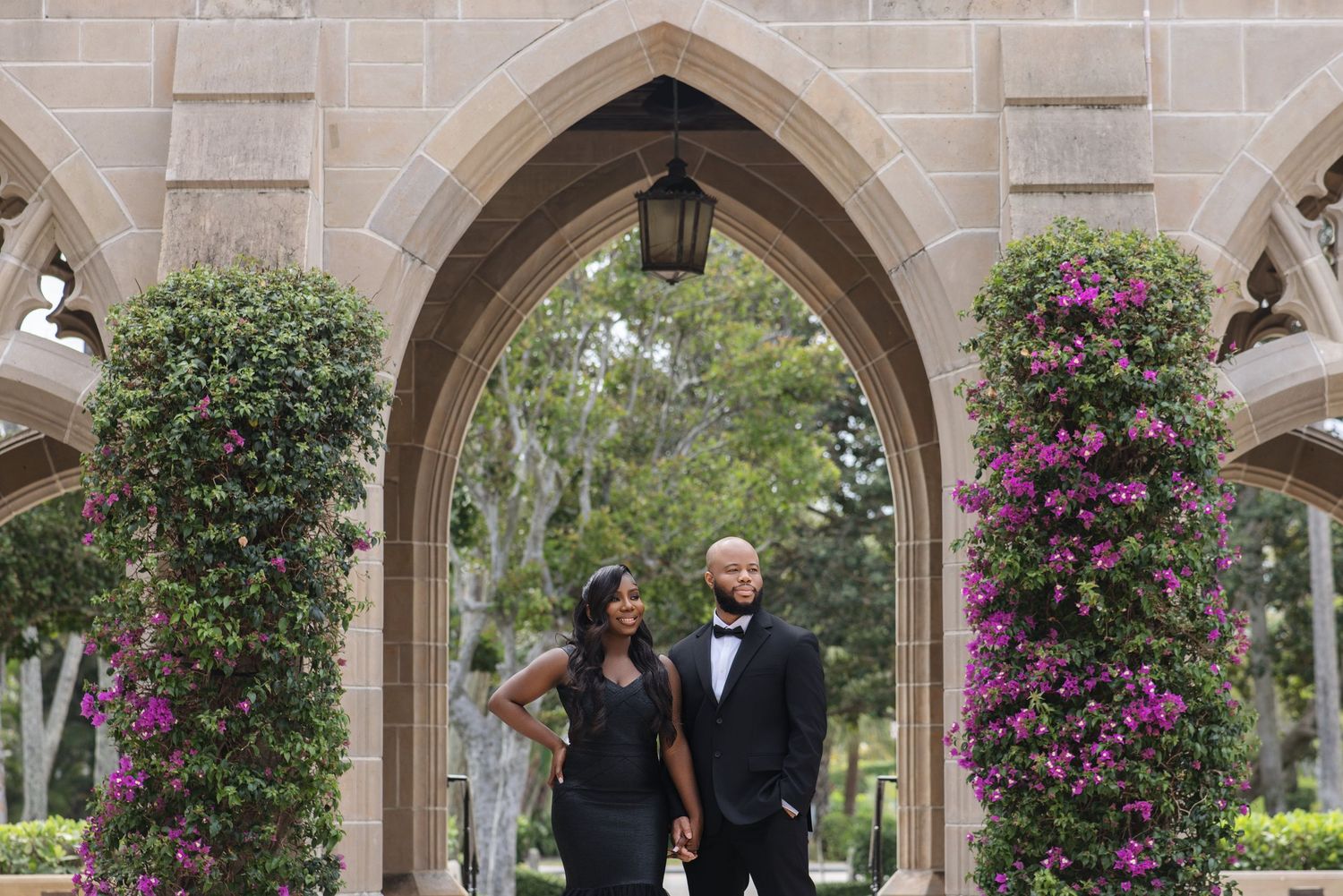Black couple holding hands under a stone archway surrounded by colorful bougainvillea blooms.