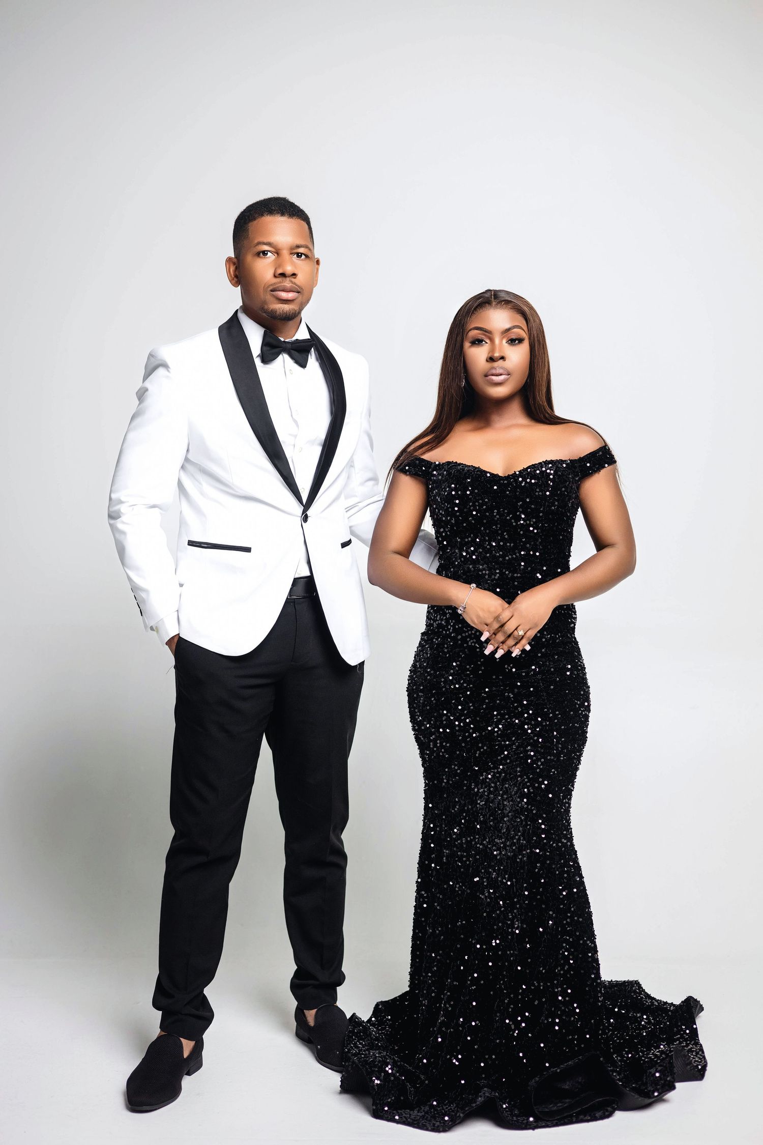Couple standing together on white backdrop wearing coordinated formal attire, black sequin gown and white tuxedo.