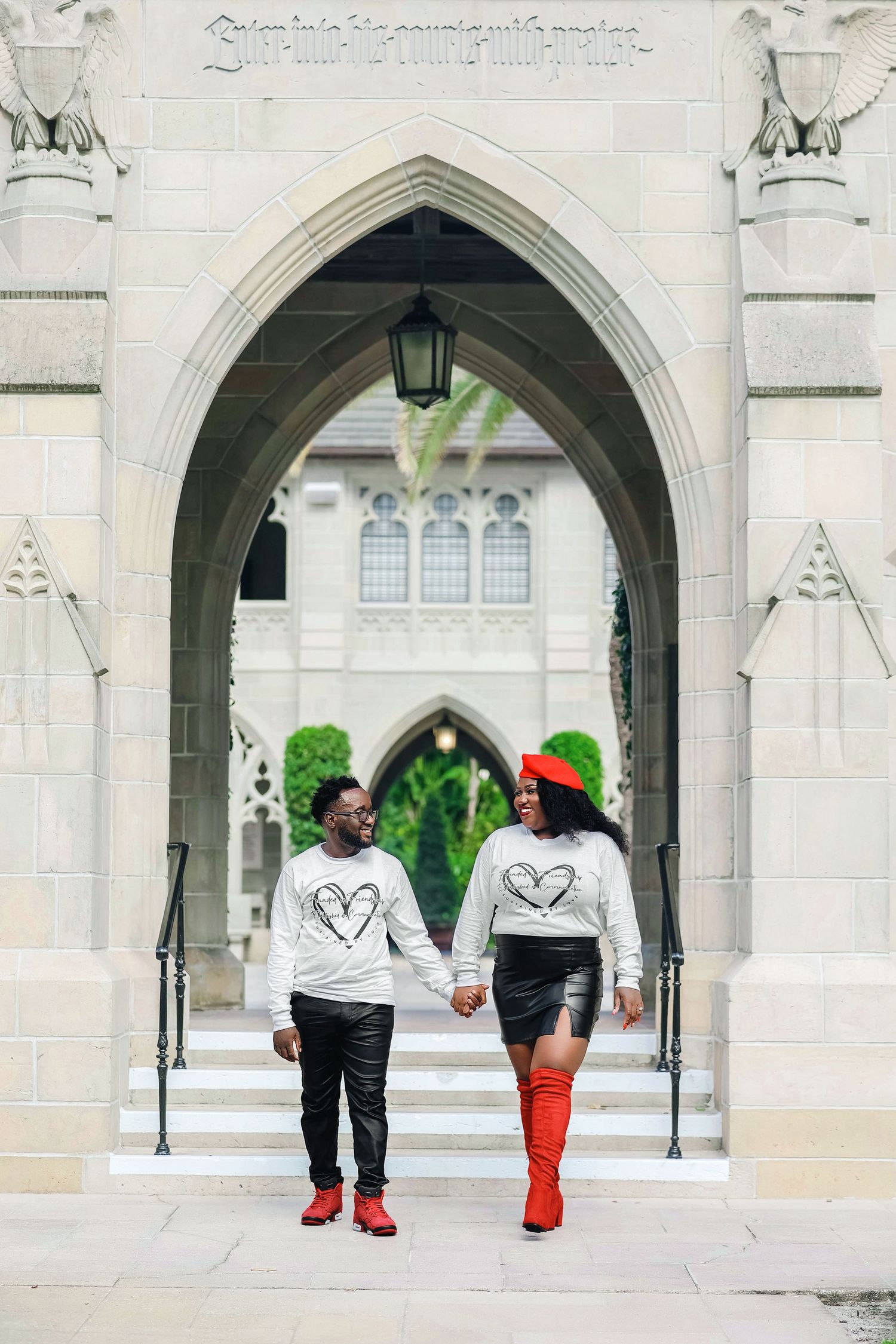 Couple in matching heart sweatshirts and red accents holding hands under a Gothic arch.