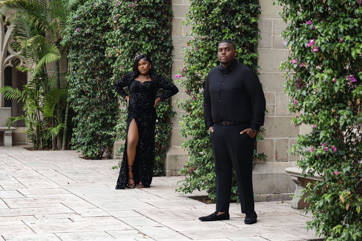 Stylish couple dressed in all-black formal attire standing in a lush garden courtyard.