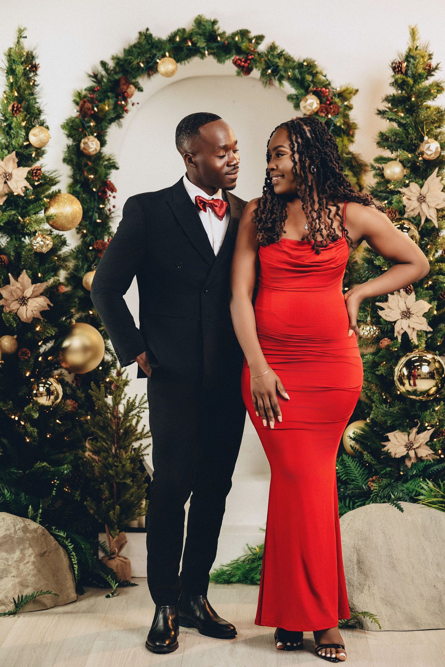Couple in red gown and black tuxedo posing before festive Christmas tree arch backdrop.