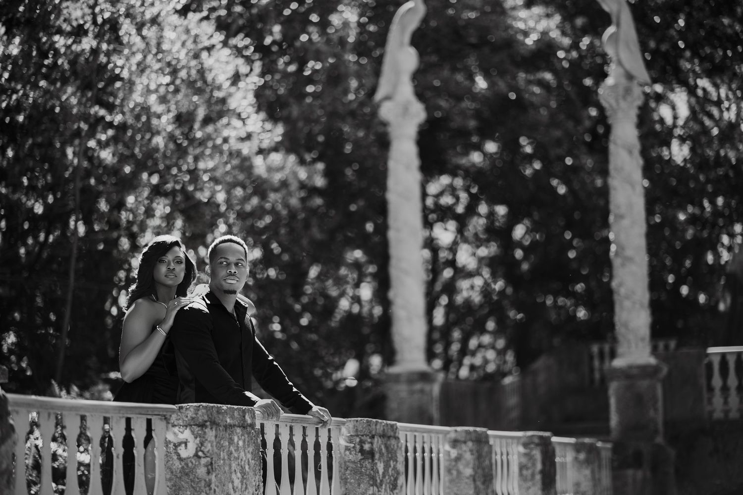 Black and white photo of couple leaning on stone balcony with garden archway behind.