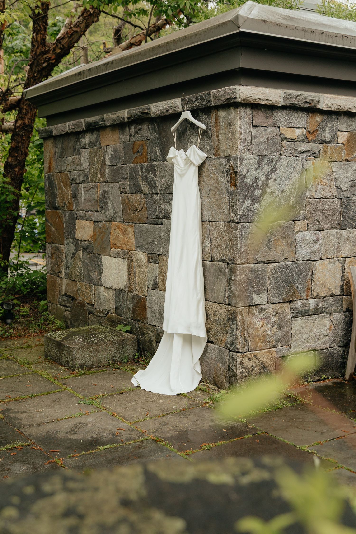 White wedding dress hanging on a stone wall outdoors surrounded by lush green trees.