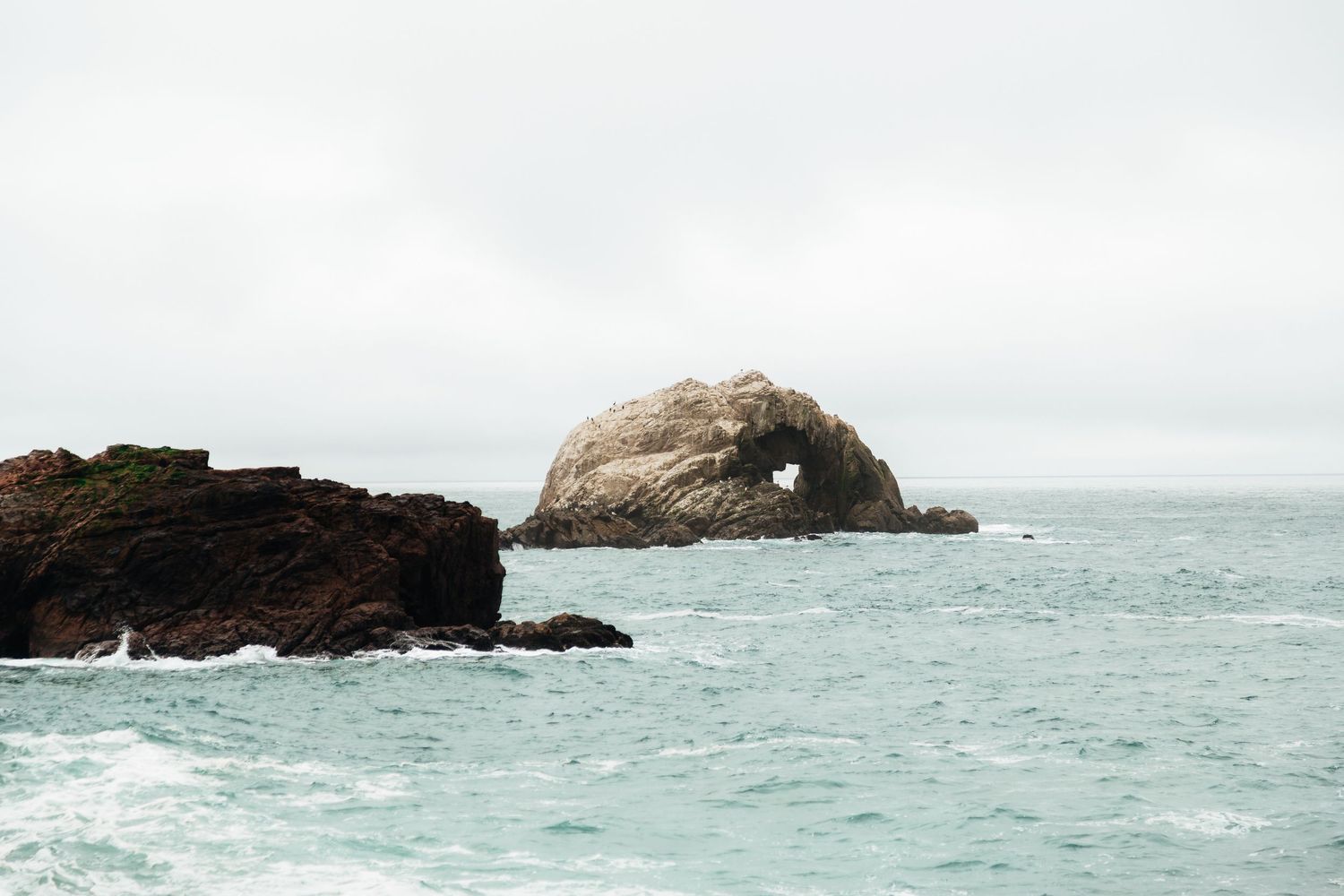A large rock formation with a natural arch stands in the calm turquoise ocean under an overcast sky.