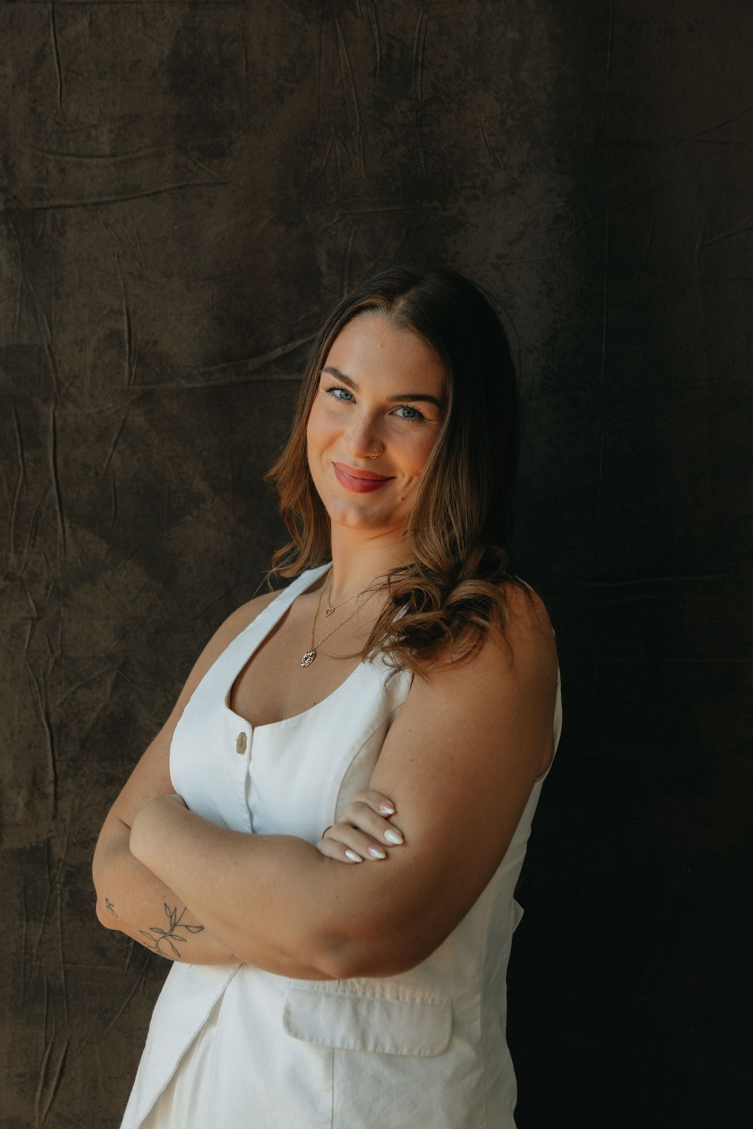 Woman in white sleeveless dress with arms crossed, smiling confidently against dark textured background.