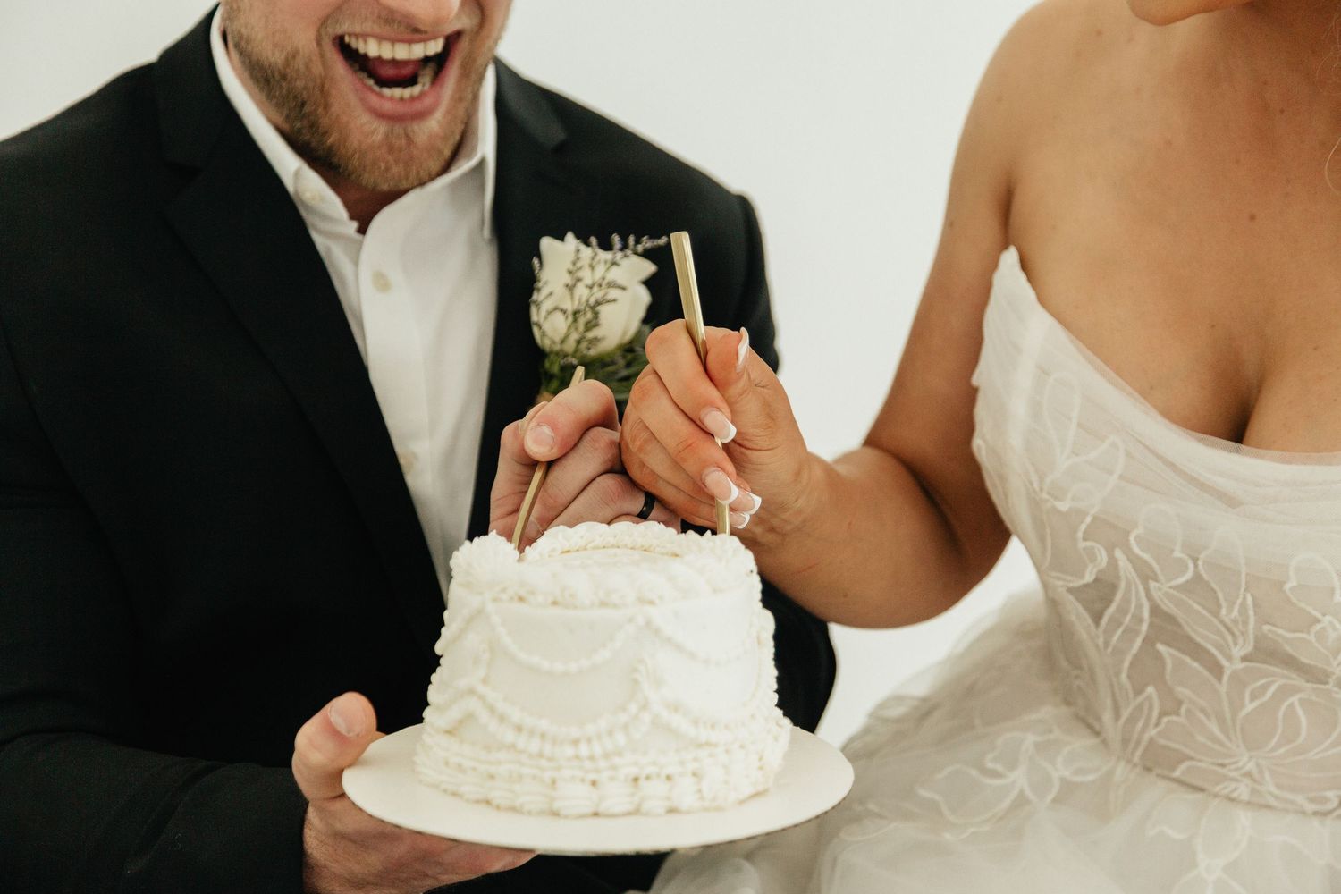 Laughing bride and groom cutting a white wedding cake with floral topper against white background.