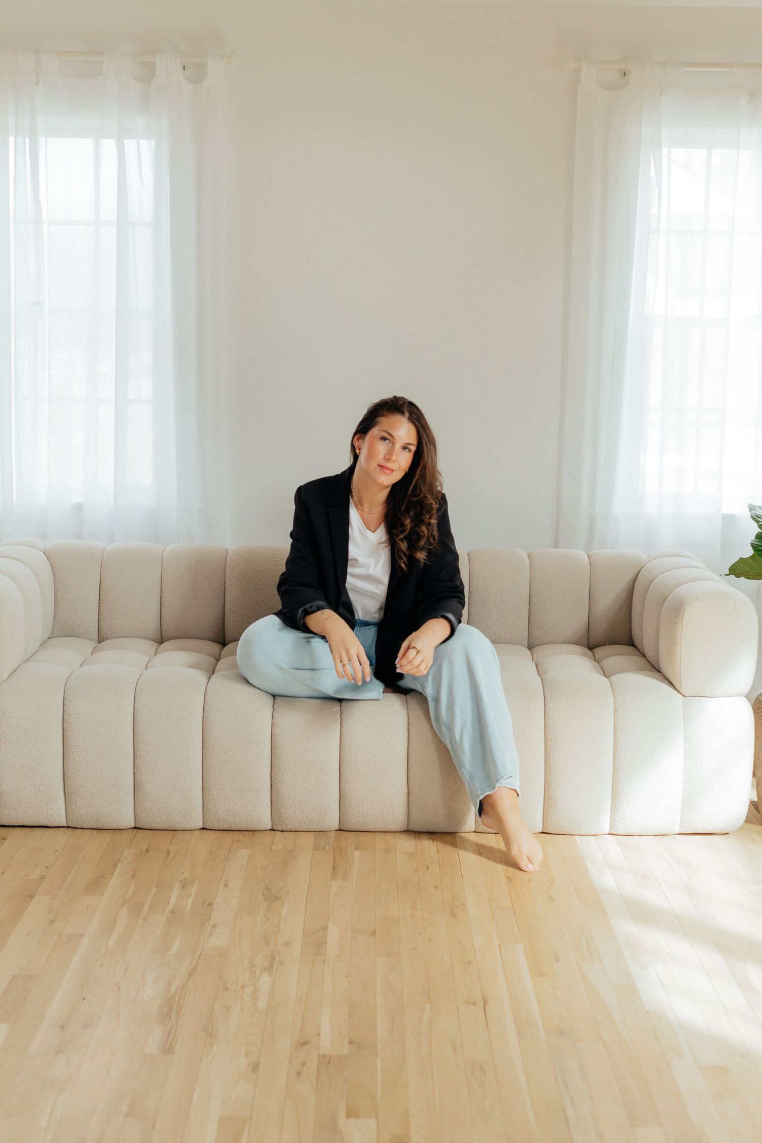 Woman in black blazer and light jeans sitting on a beige tufted sofa in a bright, minimalist living room.