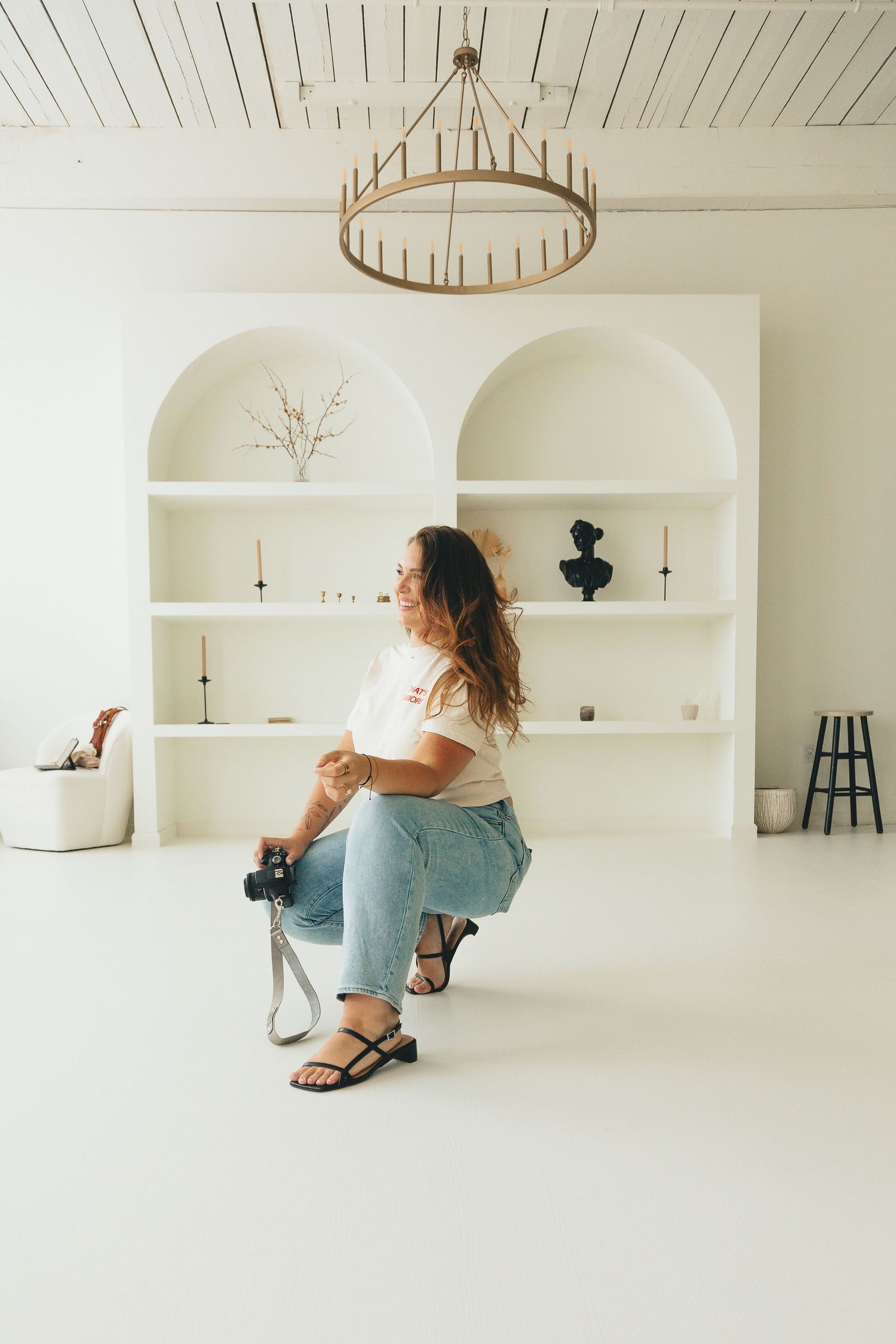 Wedding photographer sitting in a bright white studio holding a camera near arched shelving.