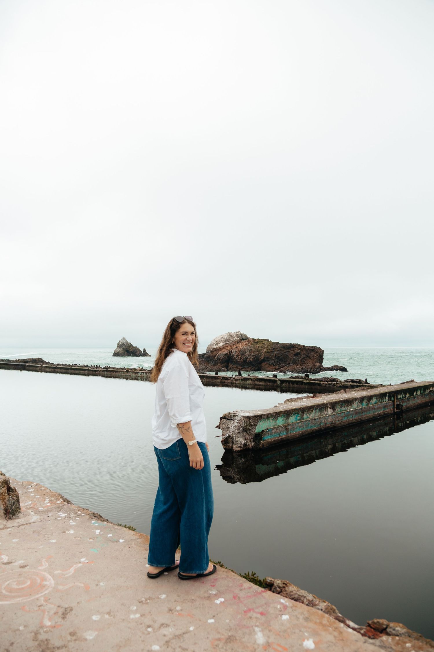 Woman in white shirt and teal wide-leg pants smiling by a coastal pier with rocky ocean views.
