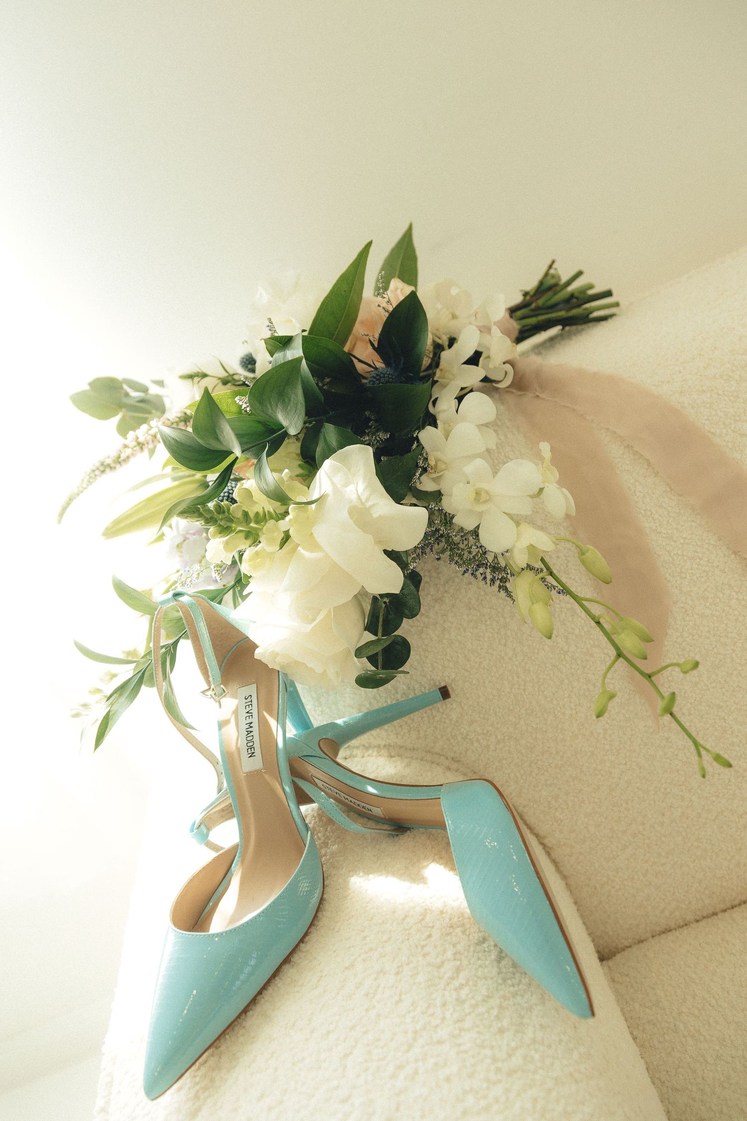Tiffany blue Jimmy Choo heels beside a white floral bridal bouquet with greenery and pink ribbon on a white chair.