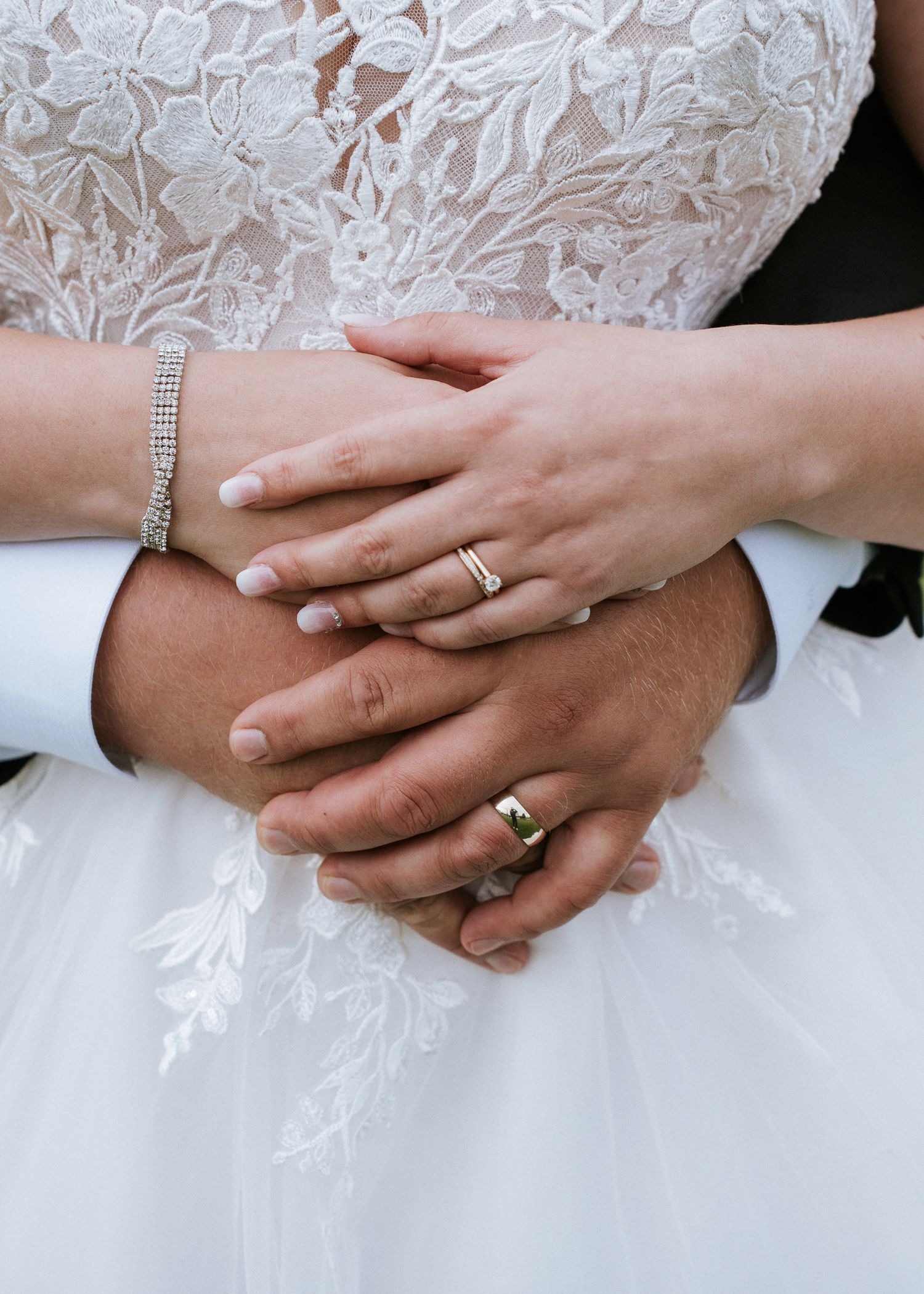 Gros plan des mains du couple de nouveaux mariés avec leurs alliances et bracelet, contre la robe de mariée en dentelle blanche.