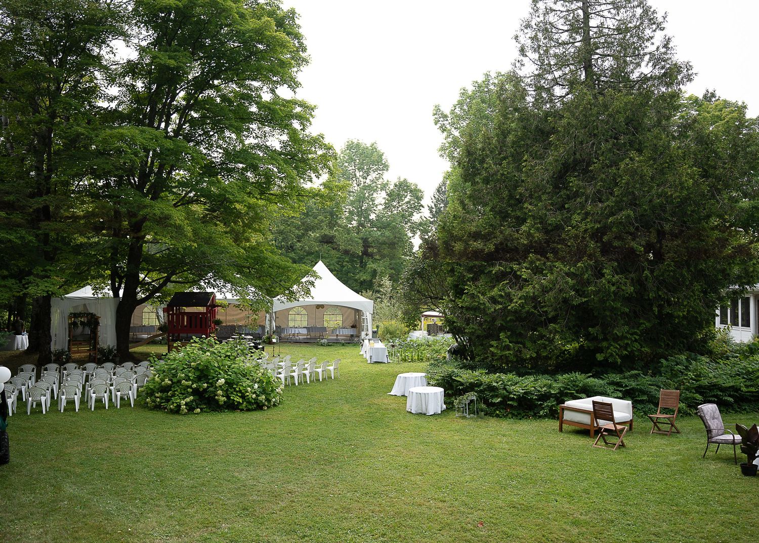 Installation de mariage en extérieur avec des chaises blanches sur une pelouse verte, des arbres et une grange rouge en arrière-plan.