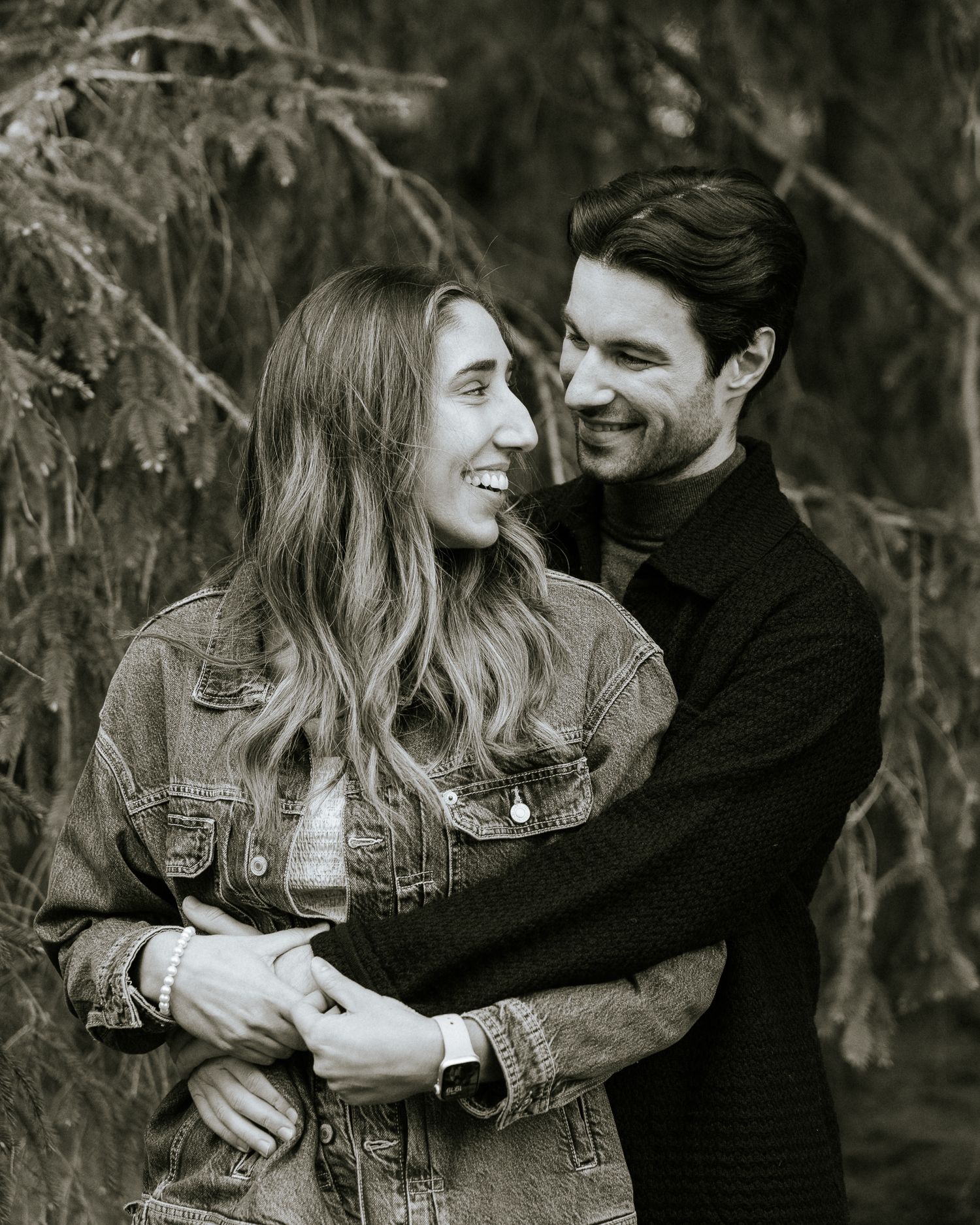 Photo spontanée en noir et blanc d'un couple s'enlaçant à l'extérieur : la femme, en veste de jean, rit alors que l'homme, en chandail.