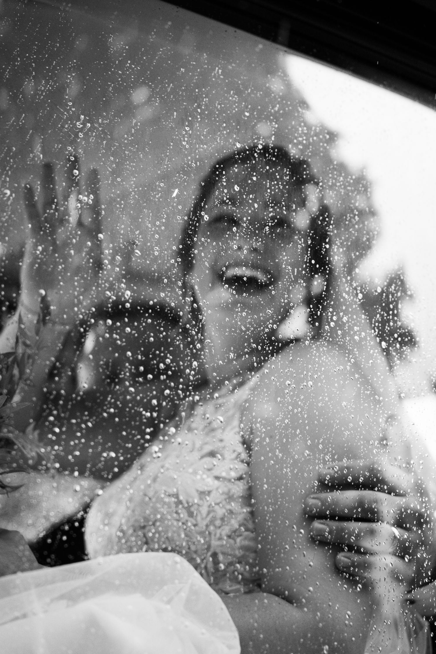 Black and white photo of a joyful bride laughing behind a rain-covered car window on her wedding day.