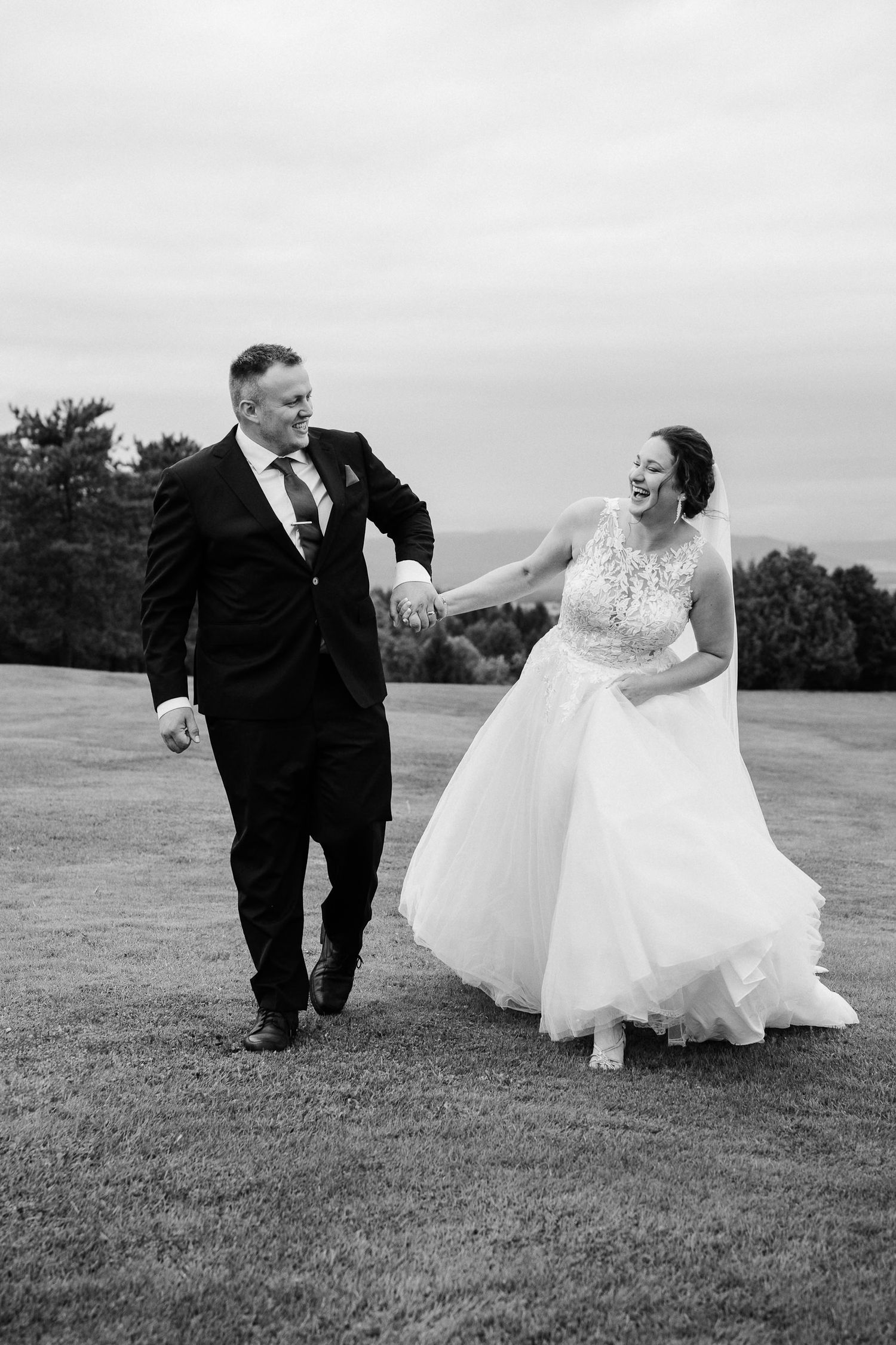 Une photo de mariage en noir et blanc capture un moment joyeux entre les nouveaux mariés marchant et riant ensemble sur une colline herbeuse
