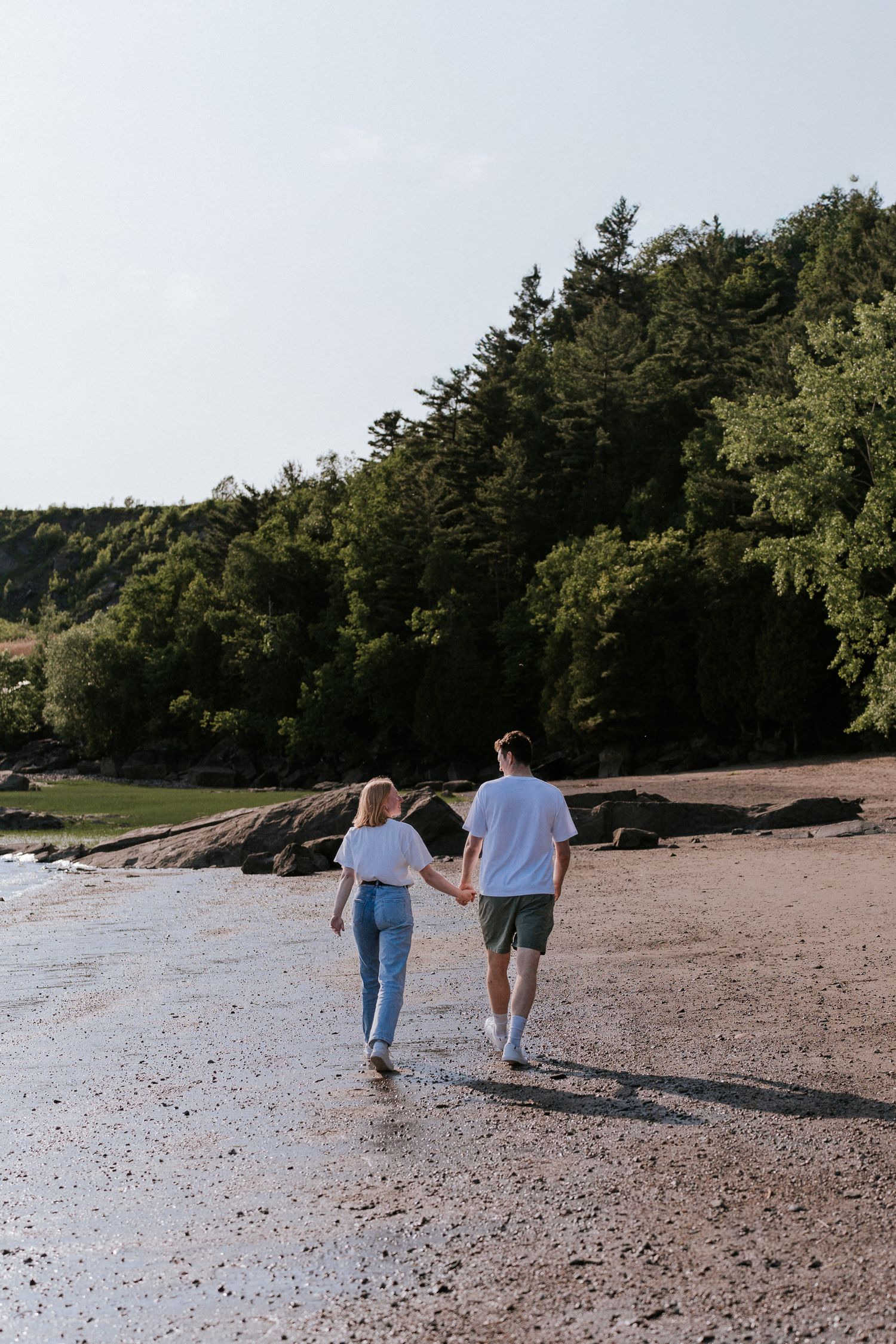 Deux personnes marchent ensemble le long d'une plage de rochers paisible, avec une forêt visible en arrière-plan.