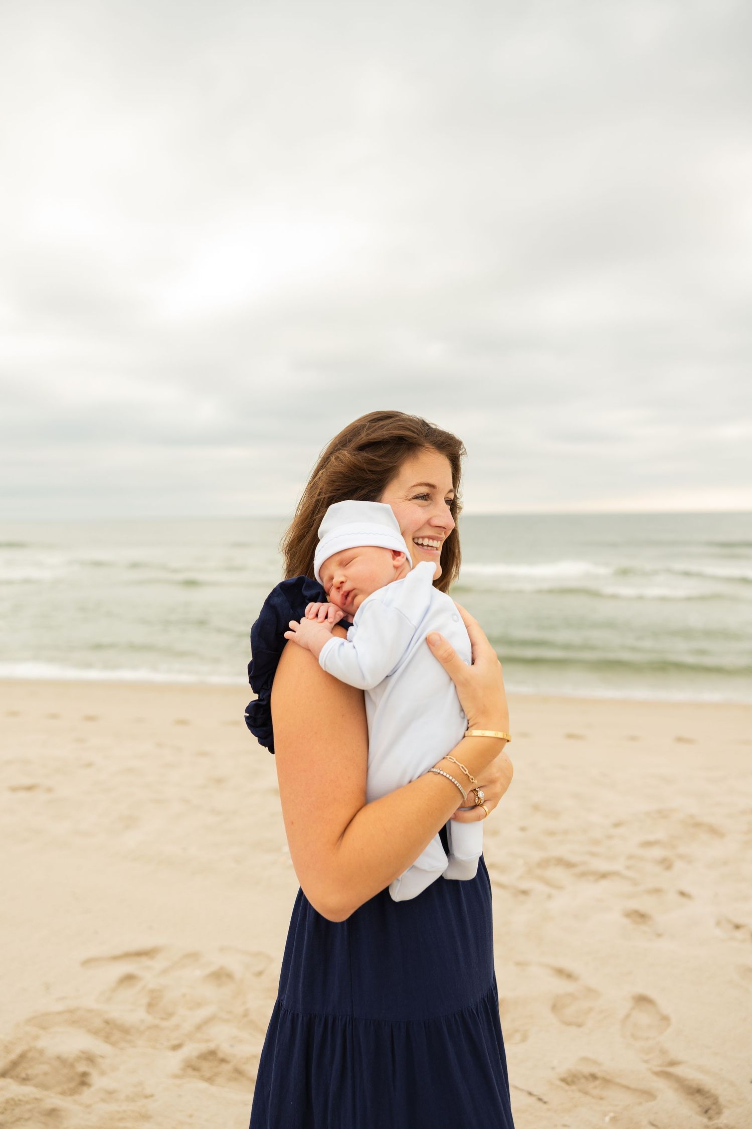 Mother and baby share intimate embrace on cloudy beach with waves in background.