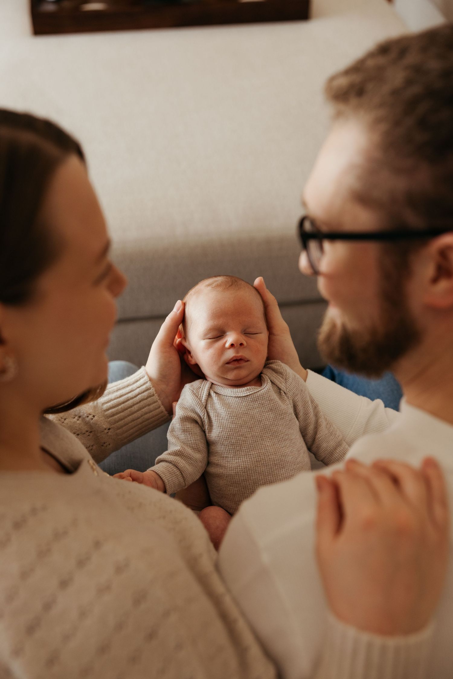 New parents tenderly cradling their sleeping newborn baby on a couch, mother gently touching infant's head.