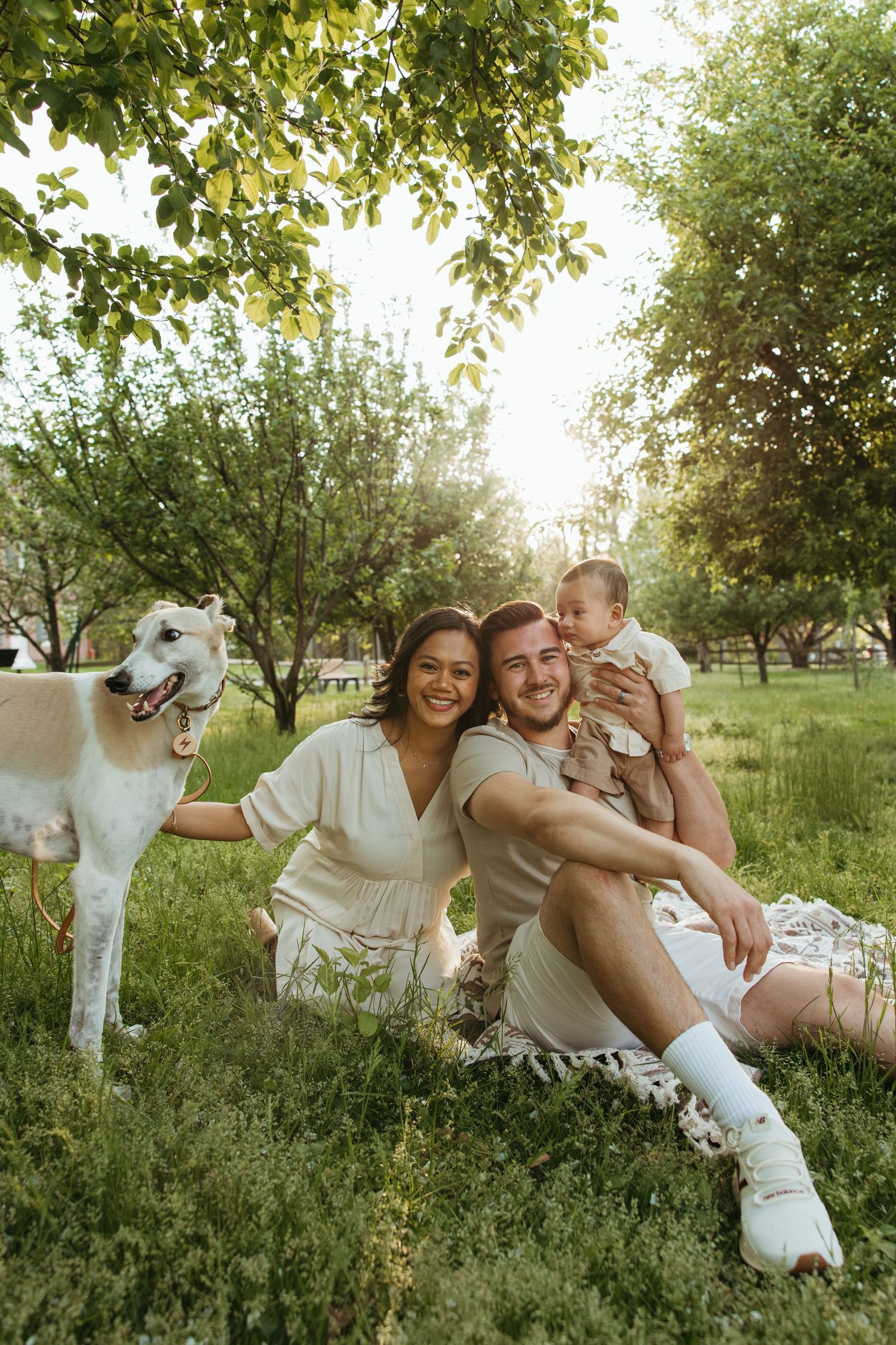 Young family with baby and white dog enjoying a sunny picnic on green grass in a lush park.