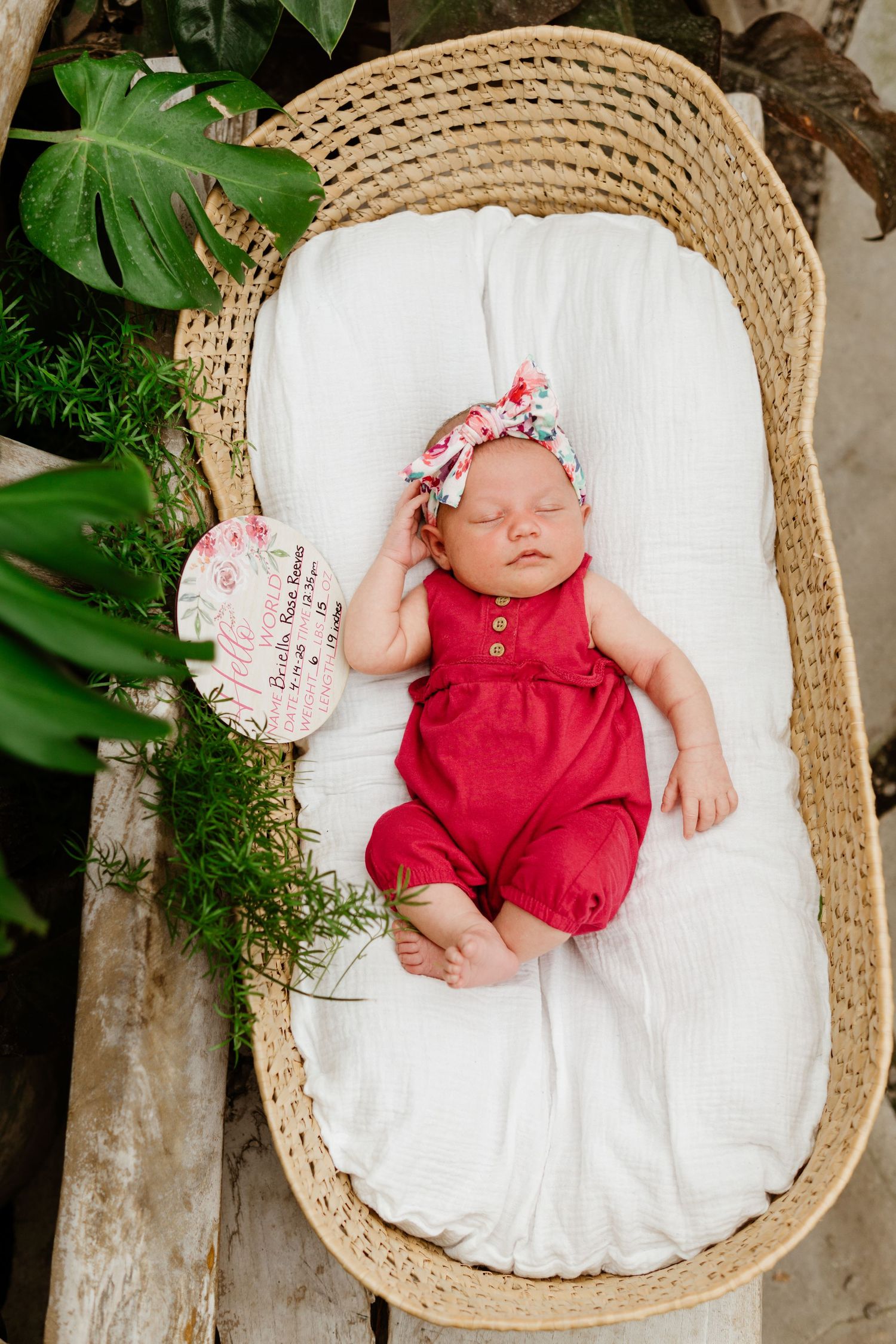 Newborn baby girl in red romper and floral headband sleeping peacefully in a wicker Moses basket with white lining.