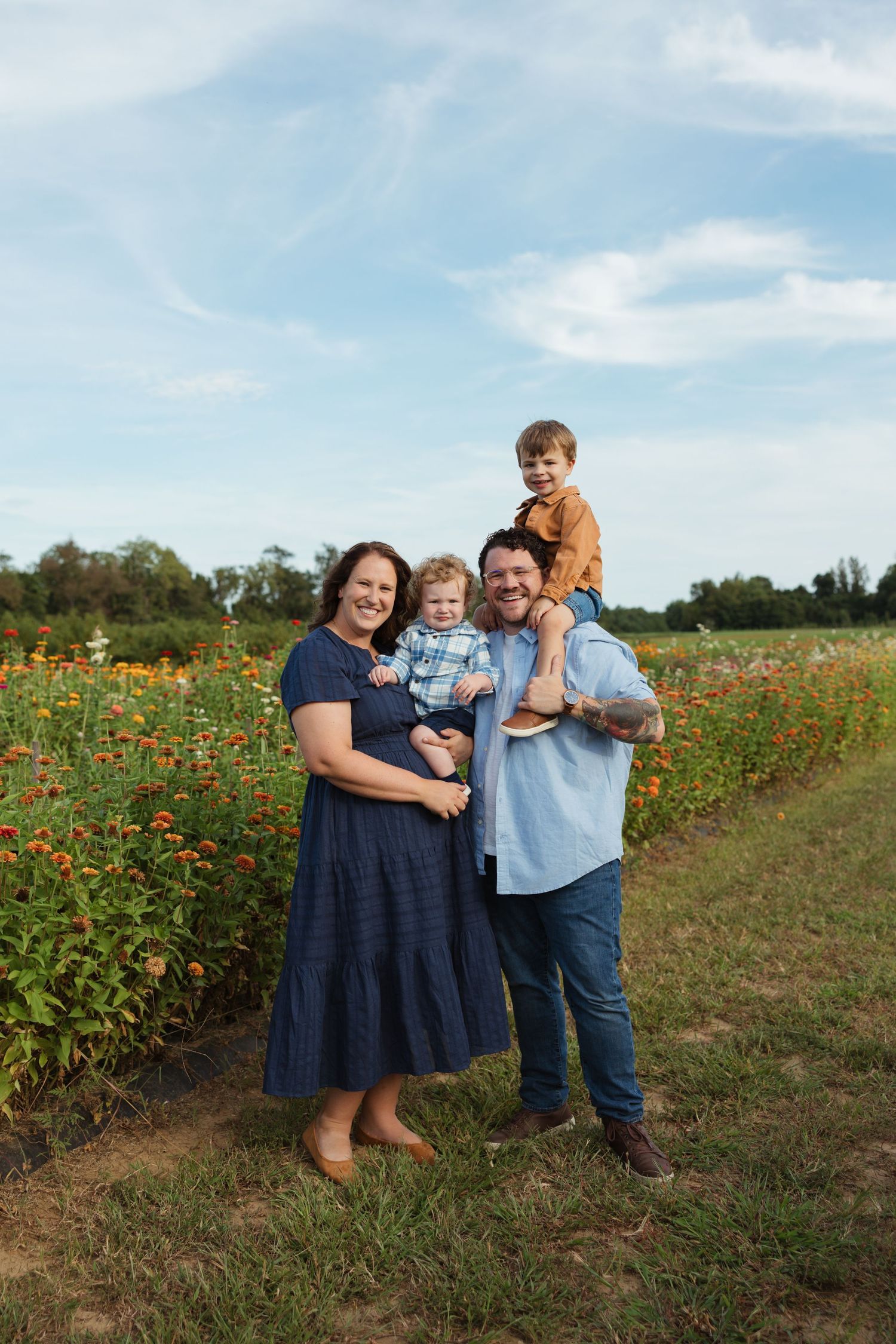 Family of four posing in a colorful flower field, parents holding two young boys, smiling outdoors under a blue sky.