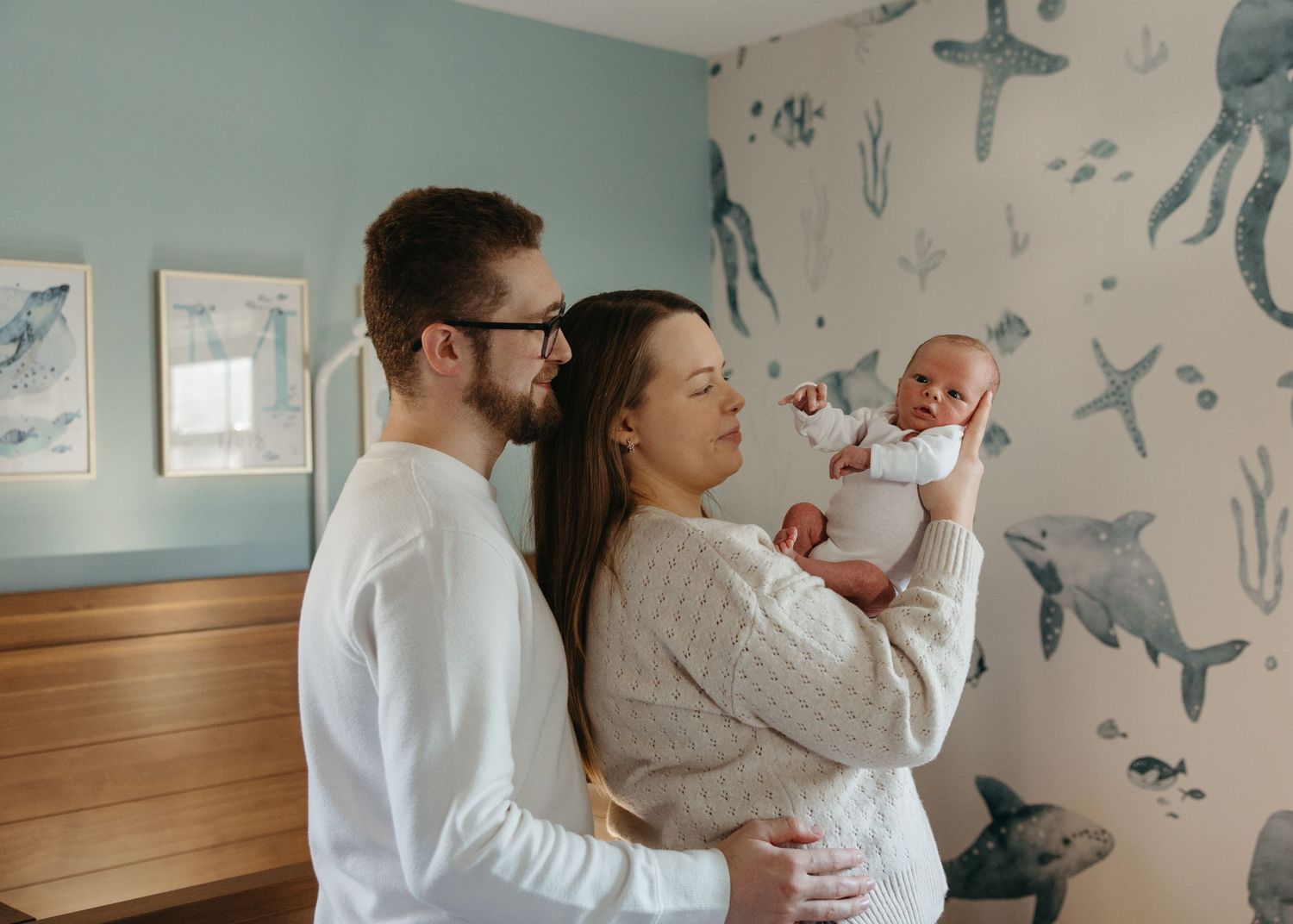 New parents admiring their newborn baby in a nursery with ocean-themed wallpaper featuring whales and sea creatures.