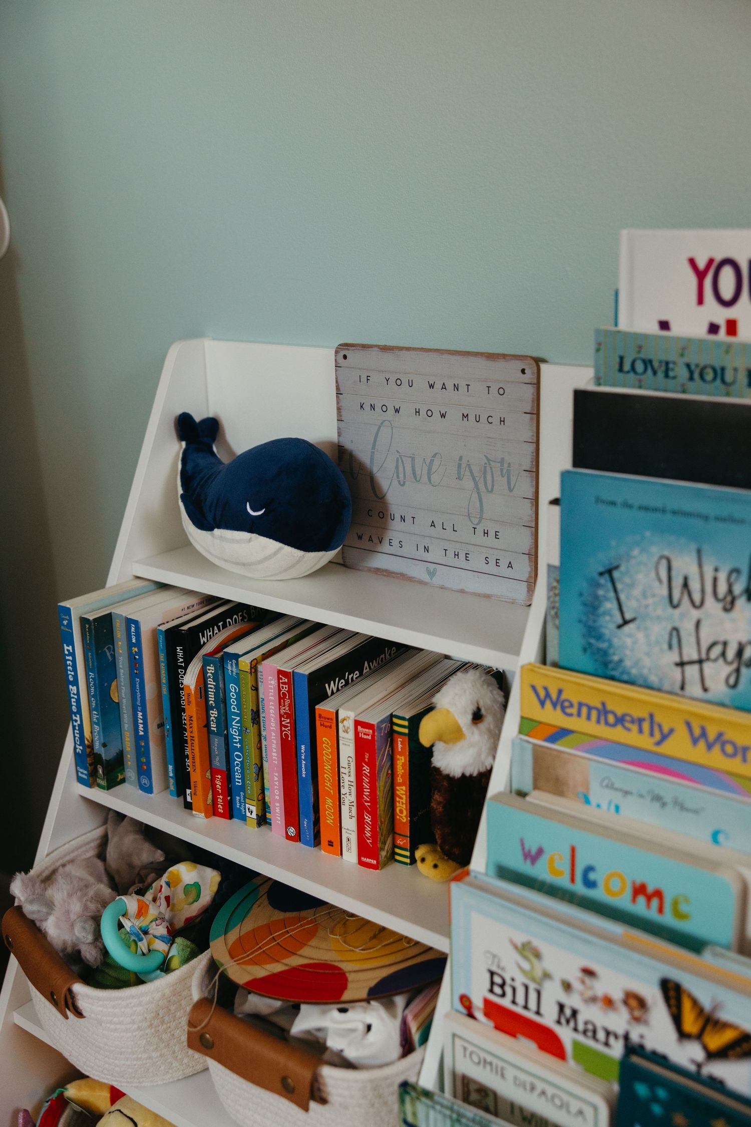 White children's bookshelf with colorful books, stuffed whale toy, eagle plush, and a welcome sign in a cozy nursery.