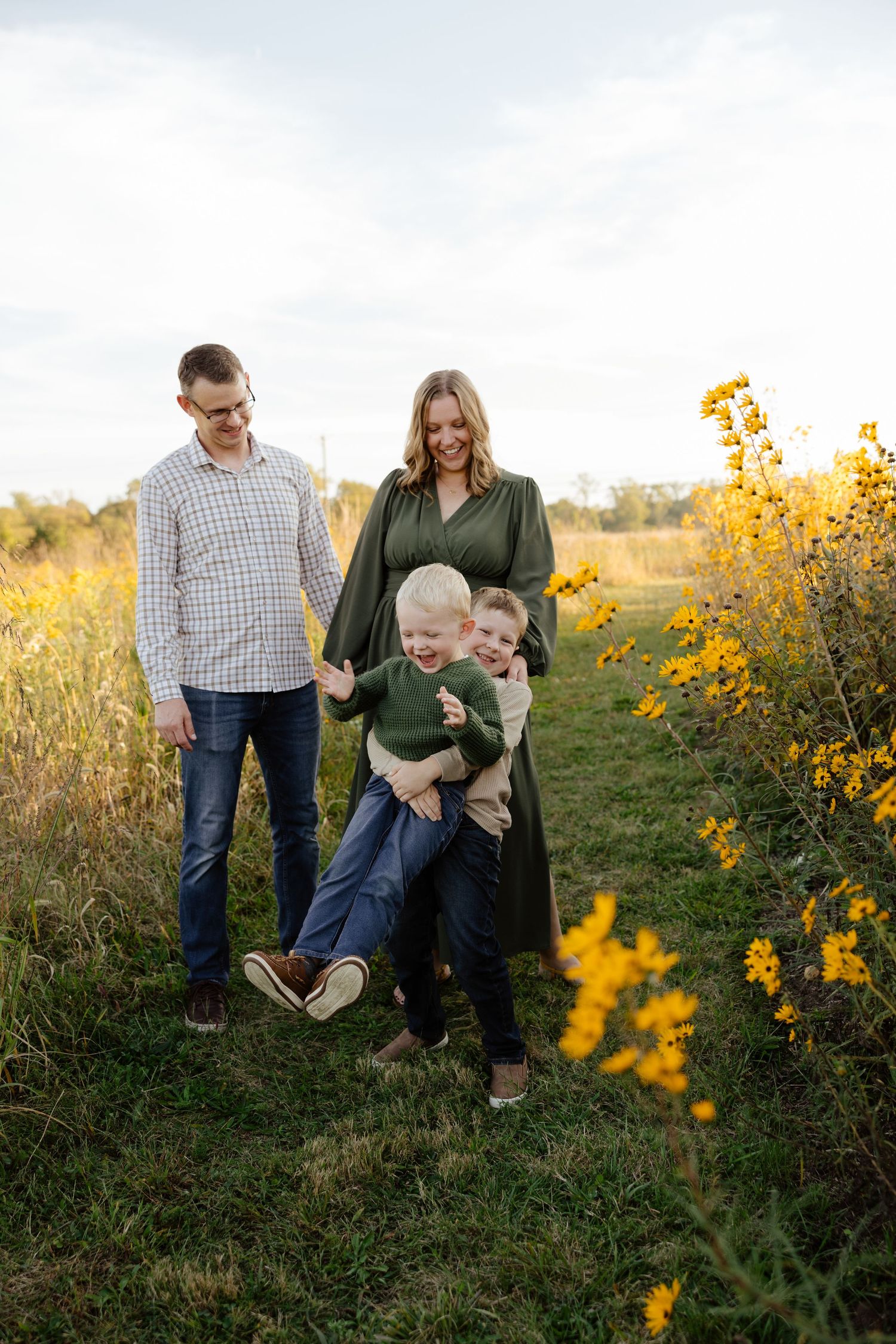 Family of four laughing together in a golden wildflower field during autumn, parents and two young boys enjoying outdoor portrait session.