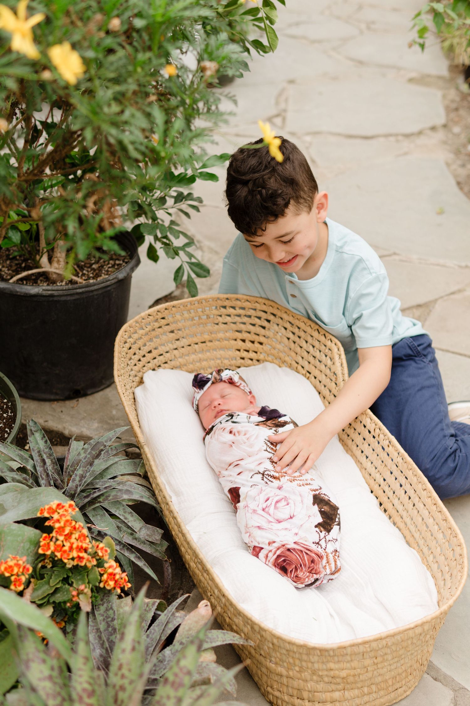 A young boy smiles while gently touching a swaddled newborn baby lying in a wicker Moses basket surrounded by plants.