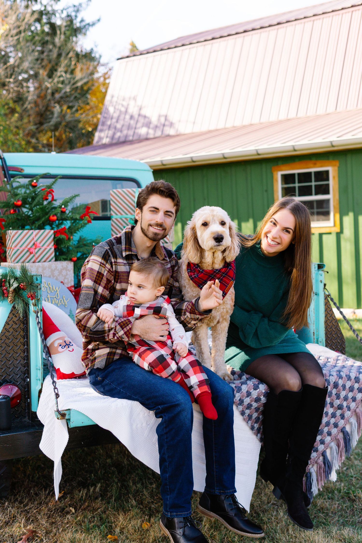 Family with baby and golden doodle dog sitting in vintage truck bed with Christmas decorations outdoors.