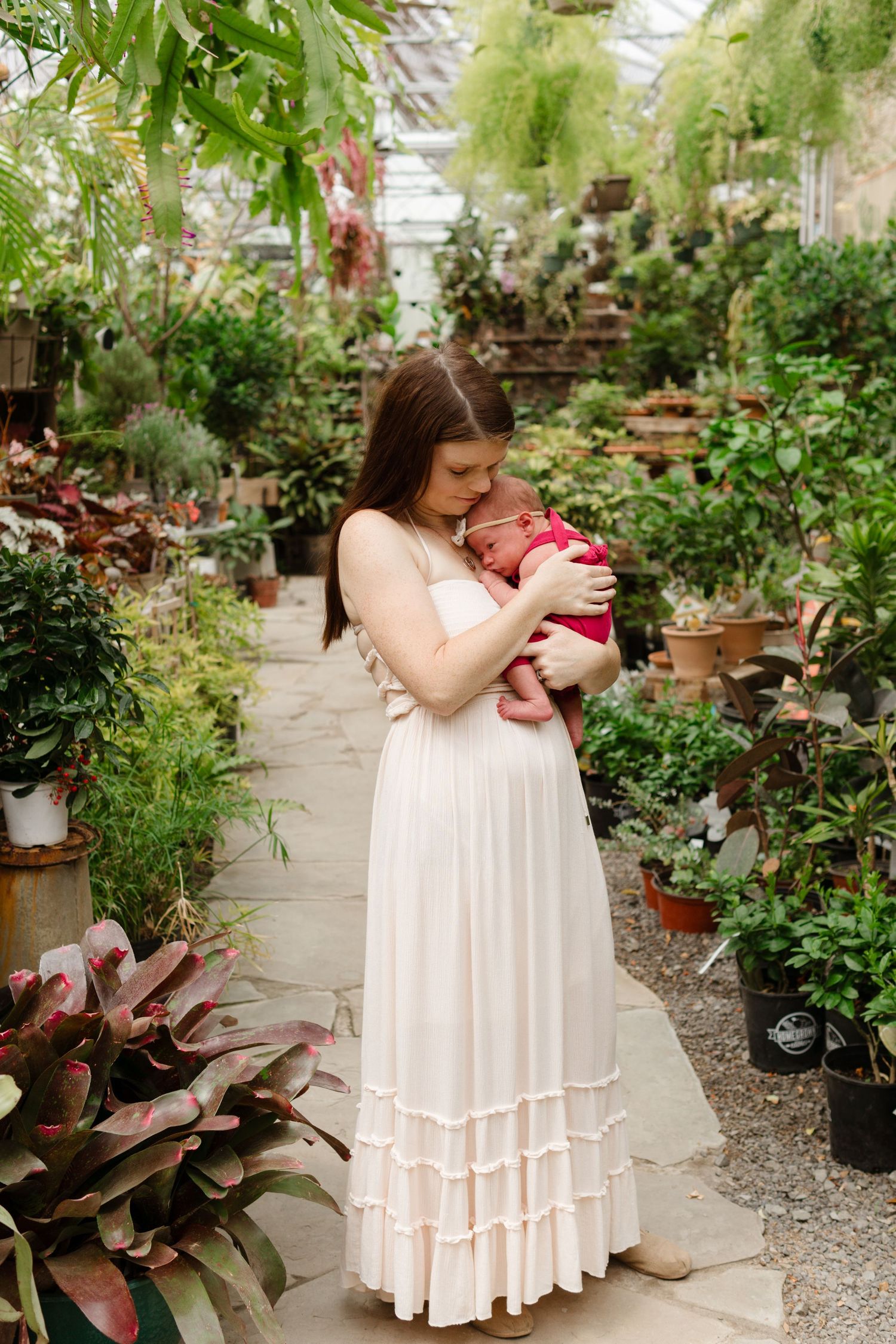 A woman in a white dress holds a newborn baby in a lush greenhouse surrounded by tropical plants.