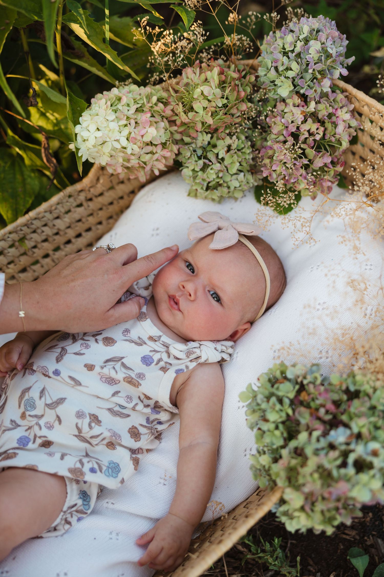 Newborn baby in floral outfit lying in wicker basket surrounded by hydrangeas with a gentle hand touching cheek.